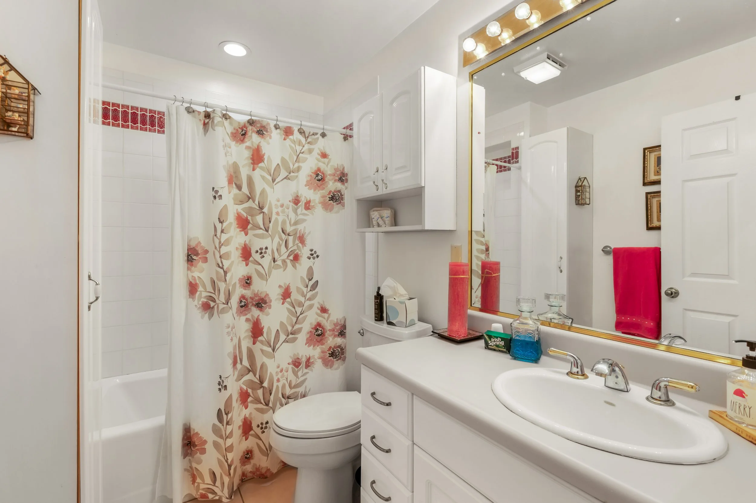 Bathroom with a floral shower curtain, white vanity with a large mirror, and various toiletries on the counter.