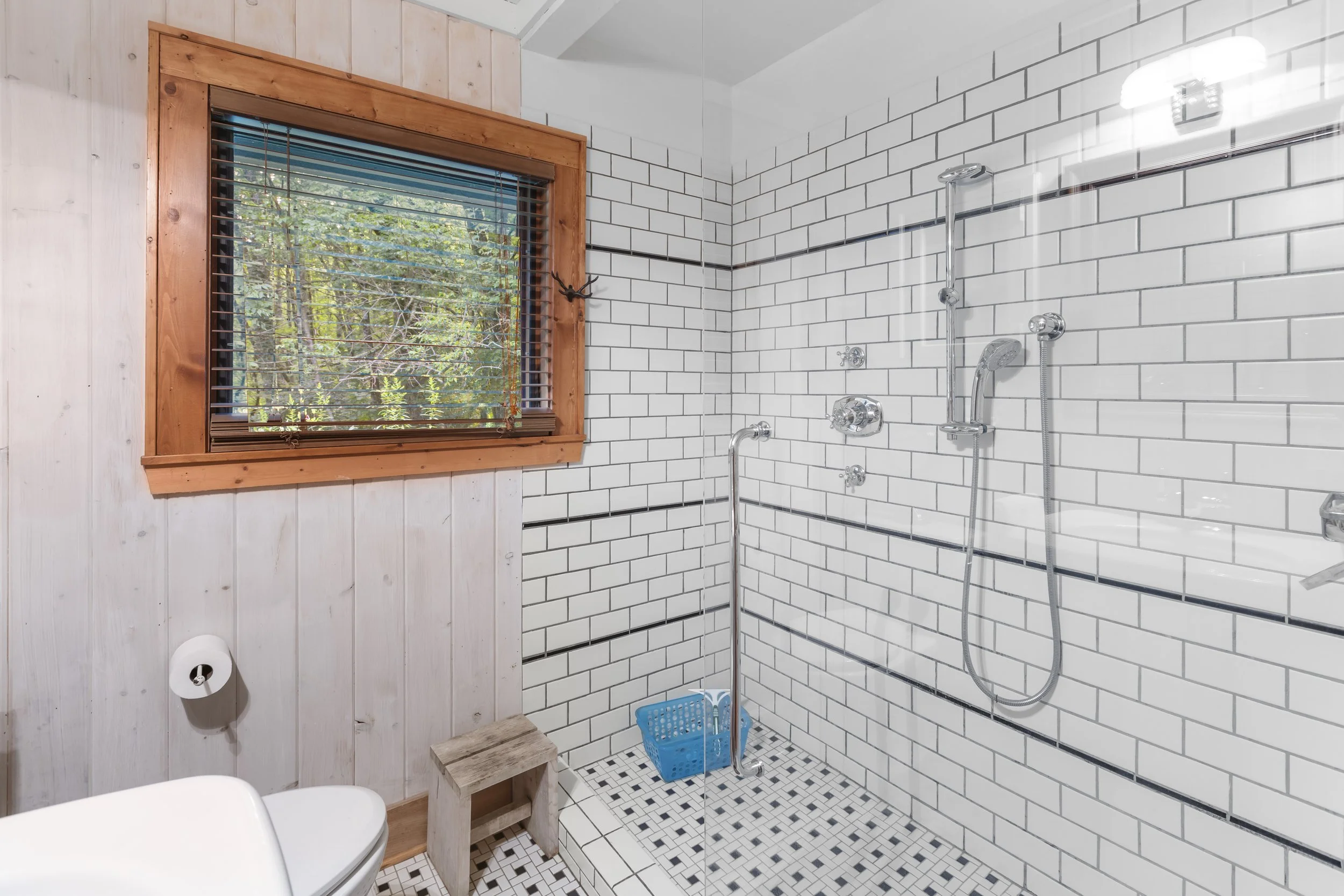Bathroom with white subway-tile wall, wooden window with blinds showing trees outside, and a walk-in shower with a blue basket on the tiled floor.