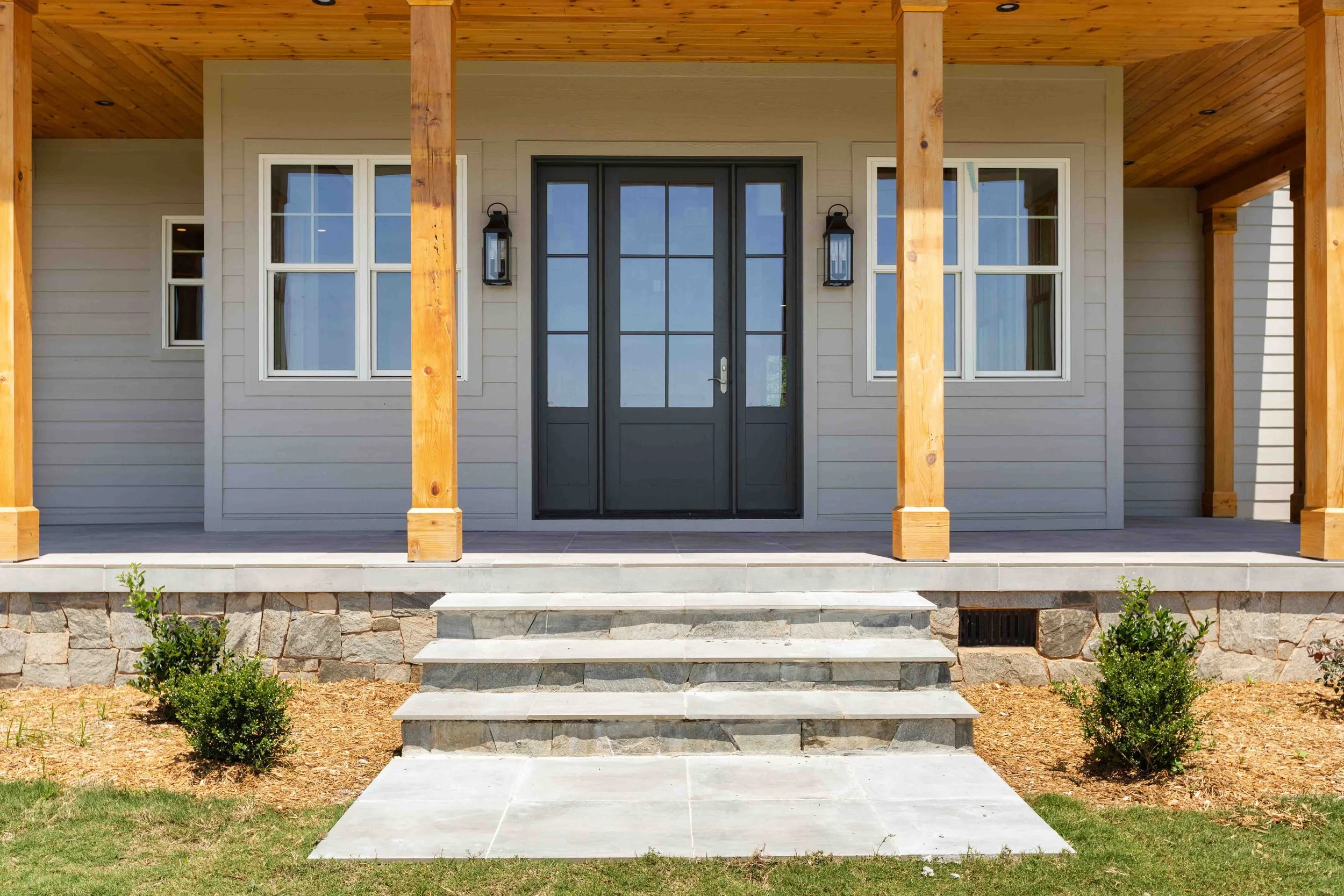 Front porch of a house with stone steps, wood-covered ceiling, gray siding, black door with glass panels, and two windows on each side of the door.