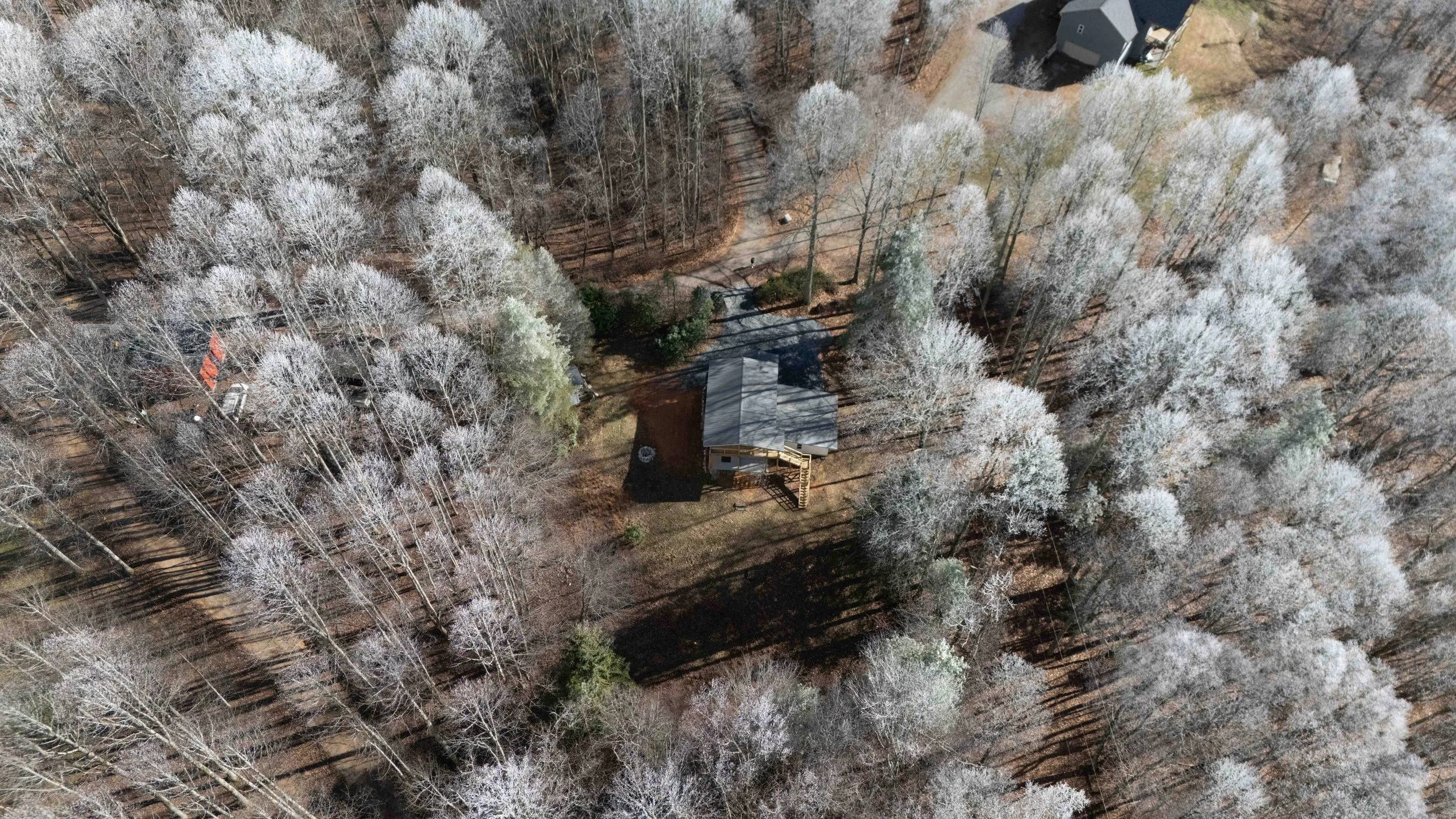 An aerial view of a house surrounded by leafless, frosty trees in a wooded area, with a driveway and nearby buildings.