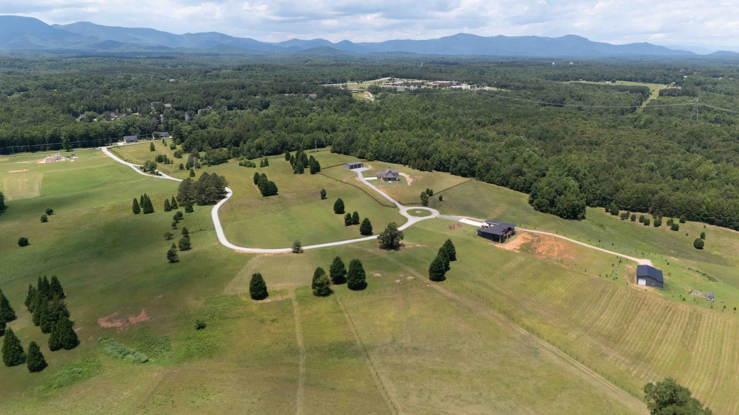 An aerial view of a rural landscape with scattered houses, winding gravel roads, green fields, and a dense forest with mountains in the background.