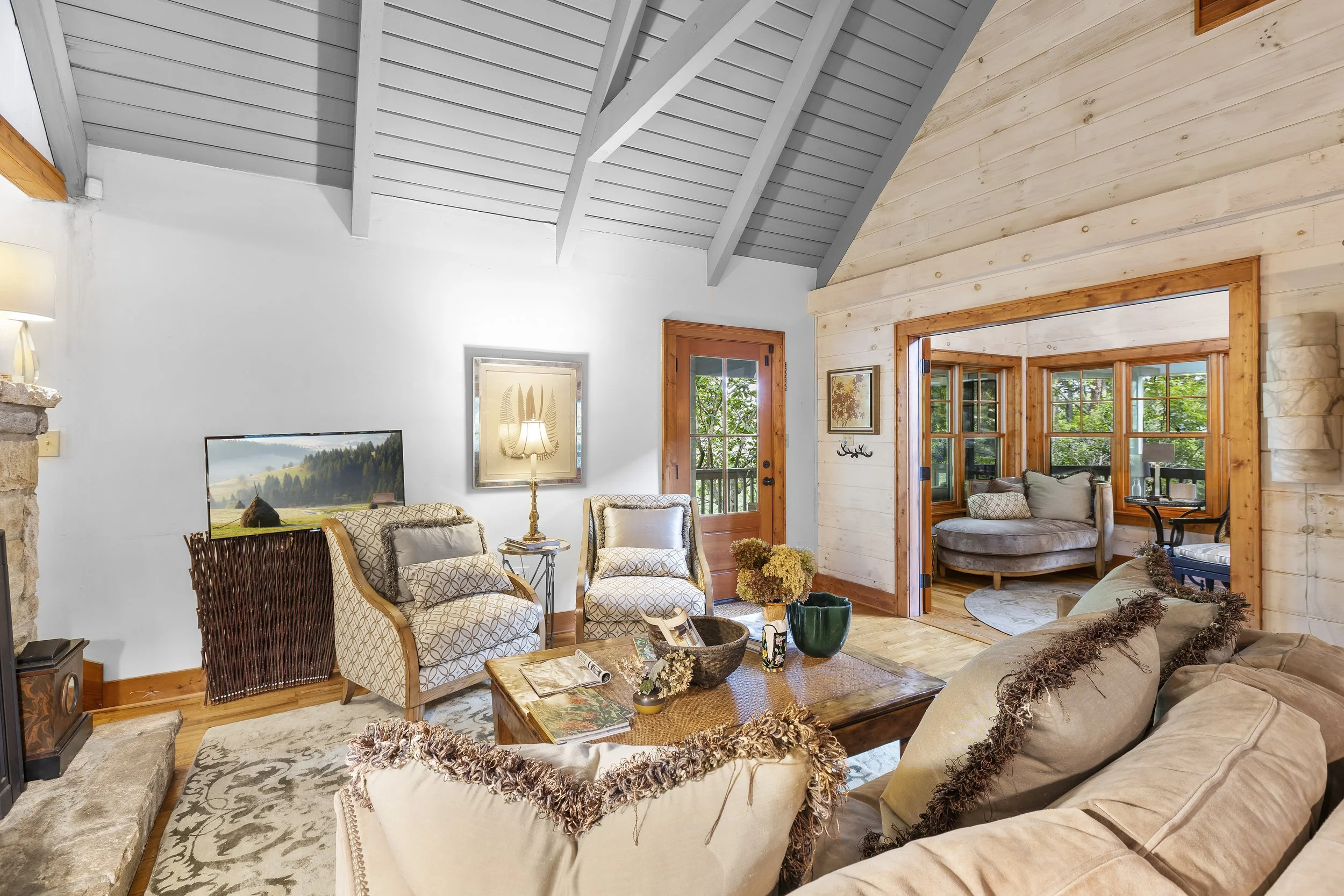 Living room with beige sofa and armchair, wooden coffee table, TV, lamps, and large windows, with a cozy, rustic decor style and a view of a green forest outside.