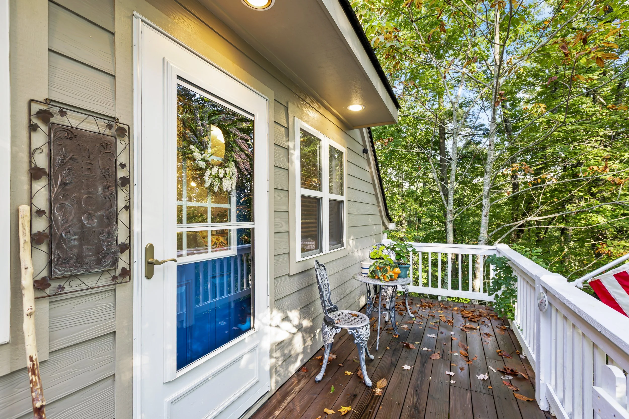 A small wooden balcony with a glass door and two windows, decorated with a metal chair, a small table with potted plants, surrounded by trees with green leaves.