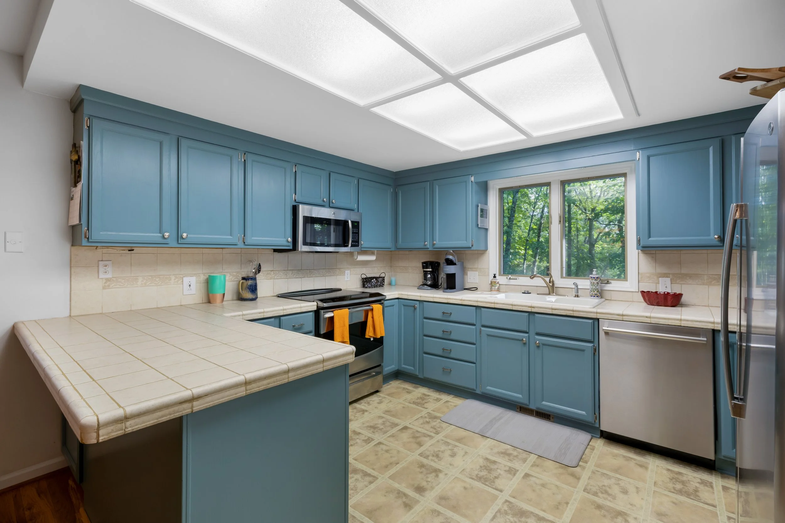 Kitchen with blue cabinets, beige tiled countertops, stainless steel appliances, and a window showing greenery outside.