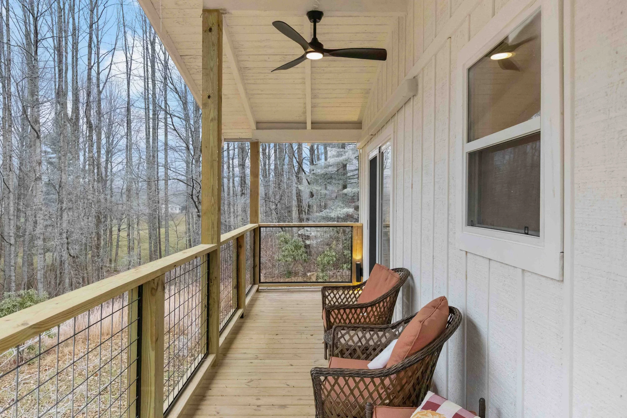 Covered balcony with wooden railing and two wicker chairs with cushions, overlooking a wooded area with leafless trees.