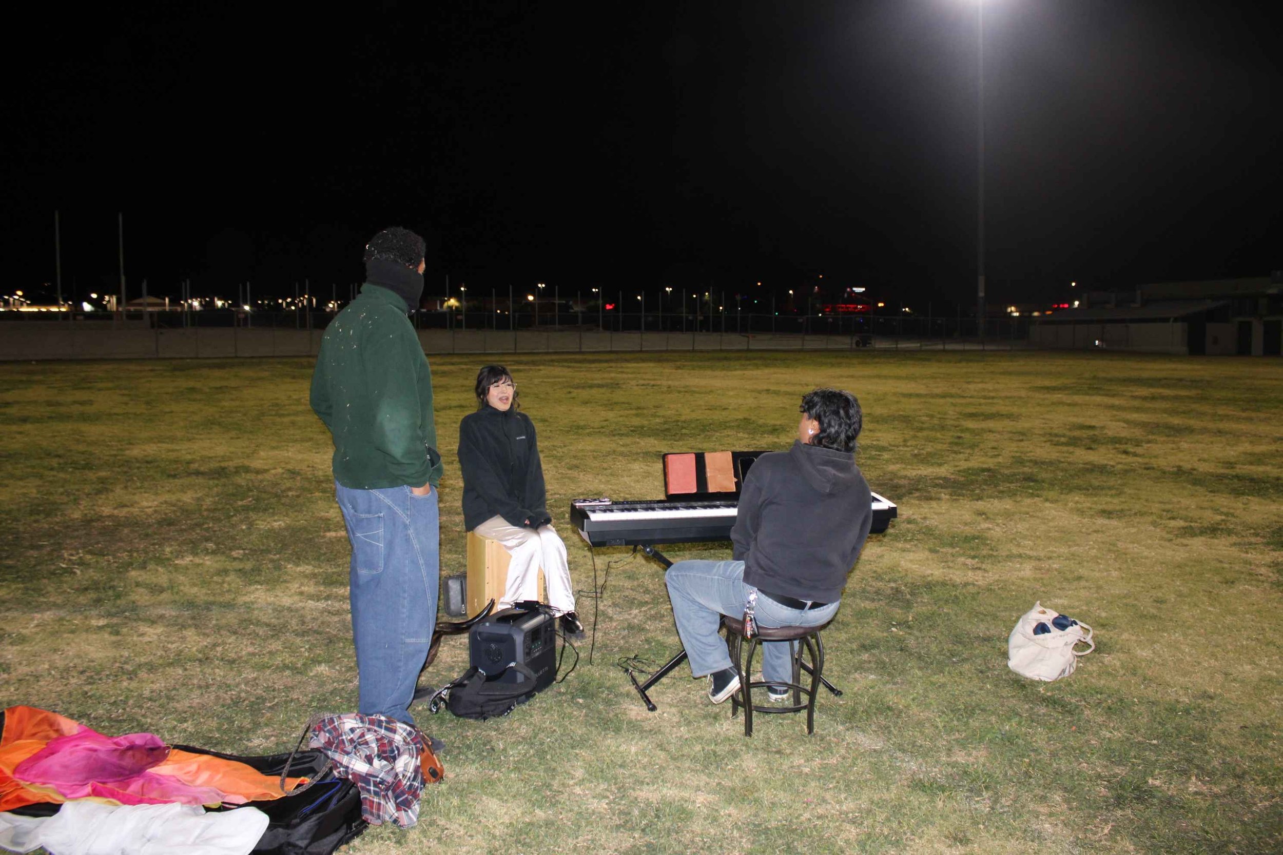 People practicing music at night on a field, featuring a keyboard and cajón.