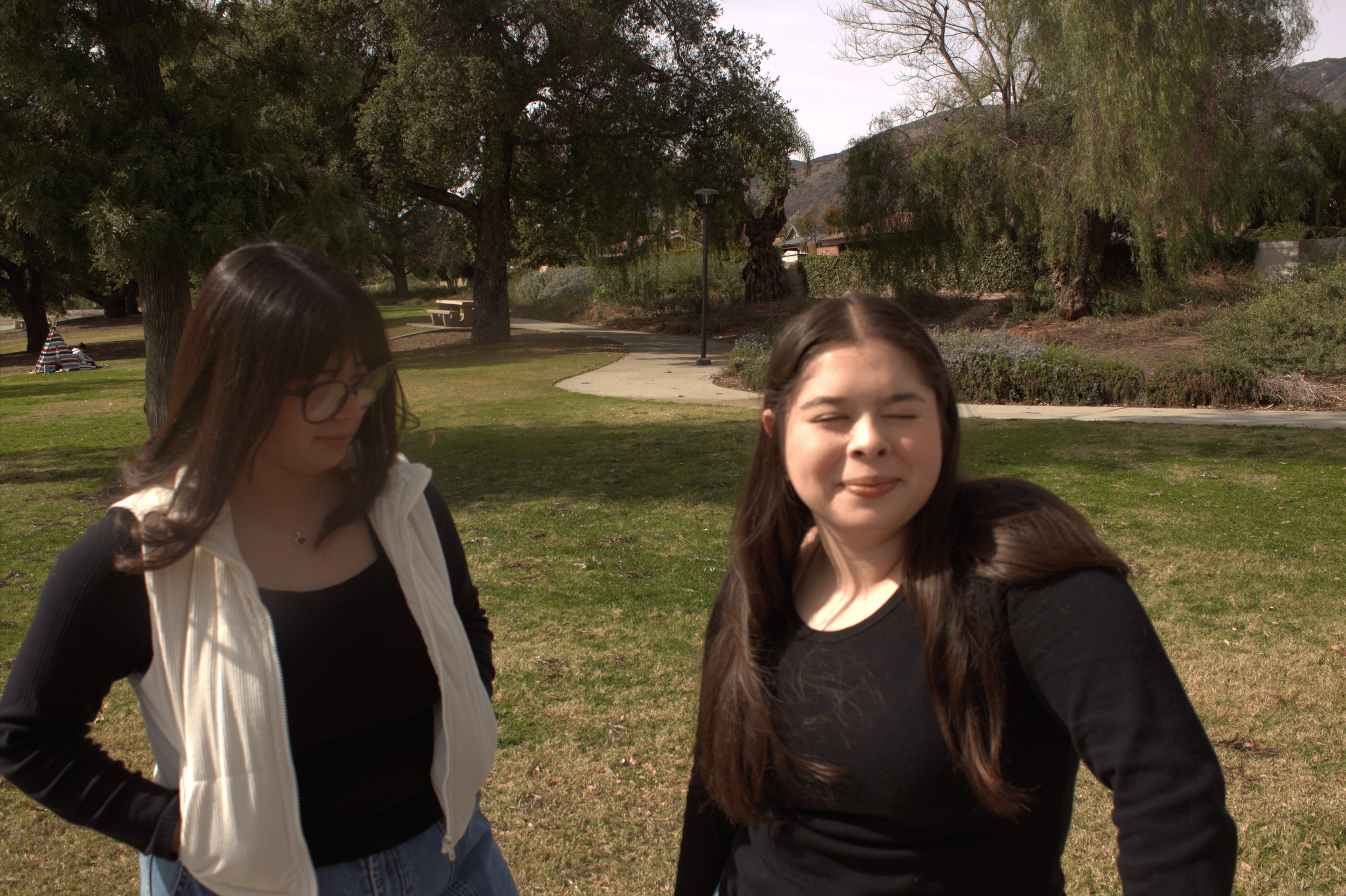 Two young women standing outdoors in a park. One has long dark hair and glasses, wearing a black top and white vest, looking down. The other has long hair, wearing a black top, with eyes closed. Trees and grass are in the background.
