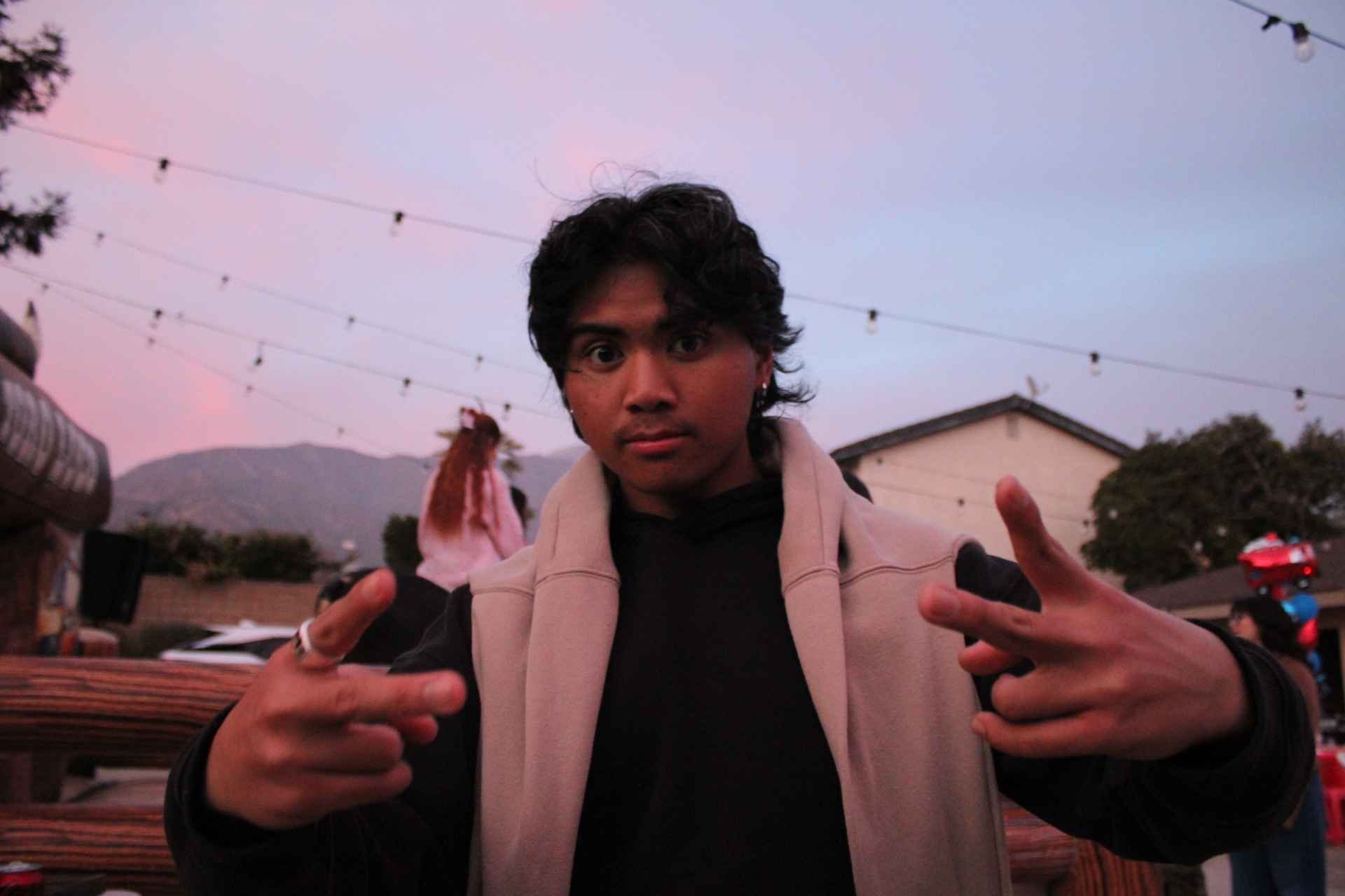 Person posing outdoors at sunset with string lights and mountains in the background.