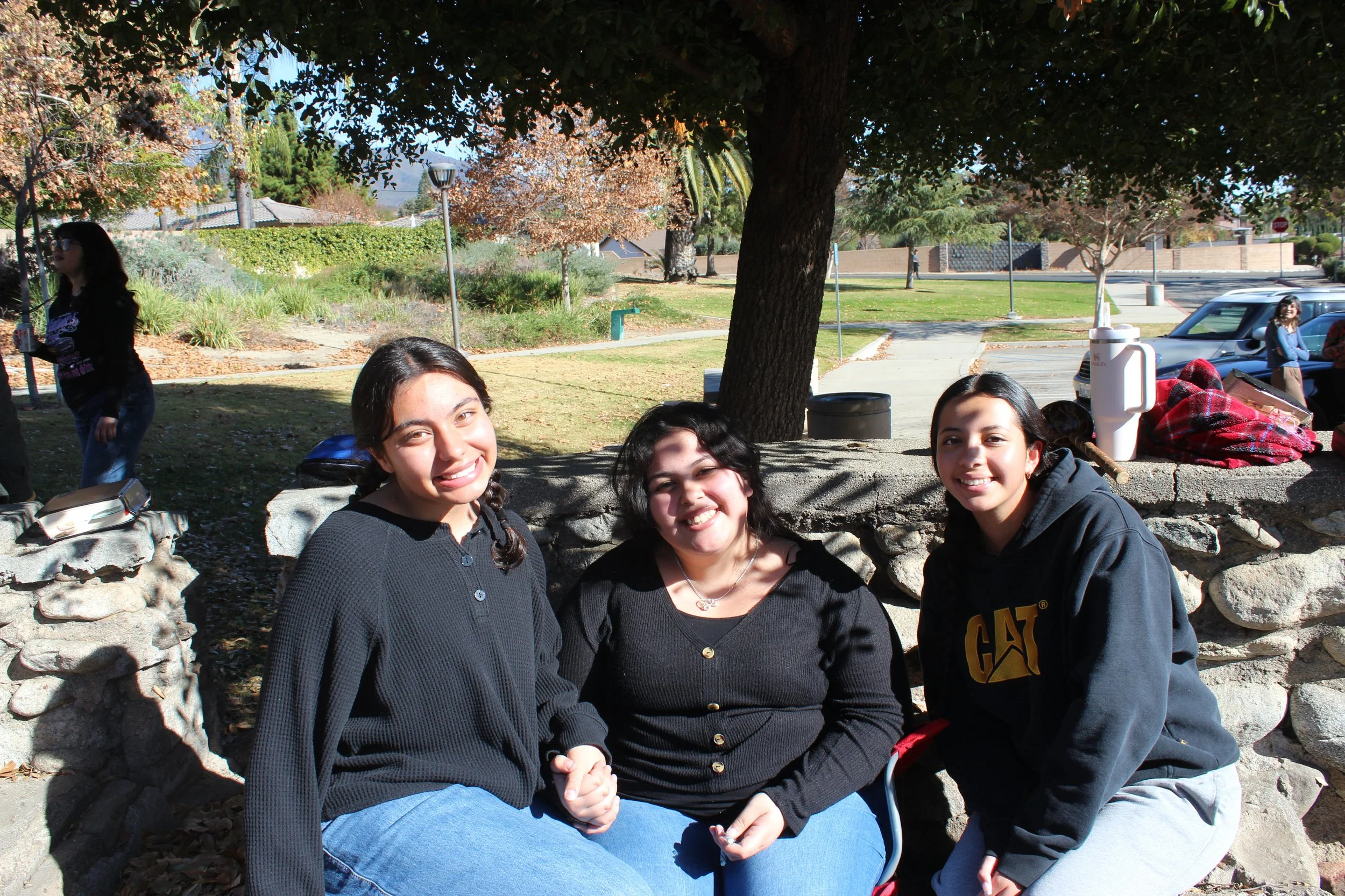Three people sitting outdoors on a stone bench, smiling. They are in a park setting with a tree providing shade. Cars and other people are visible in the background.