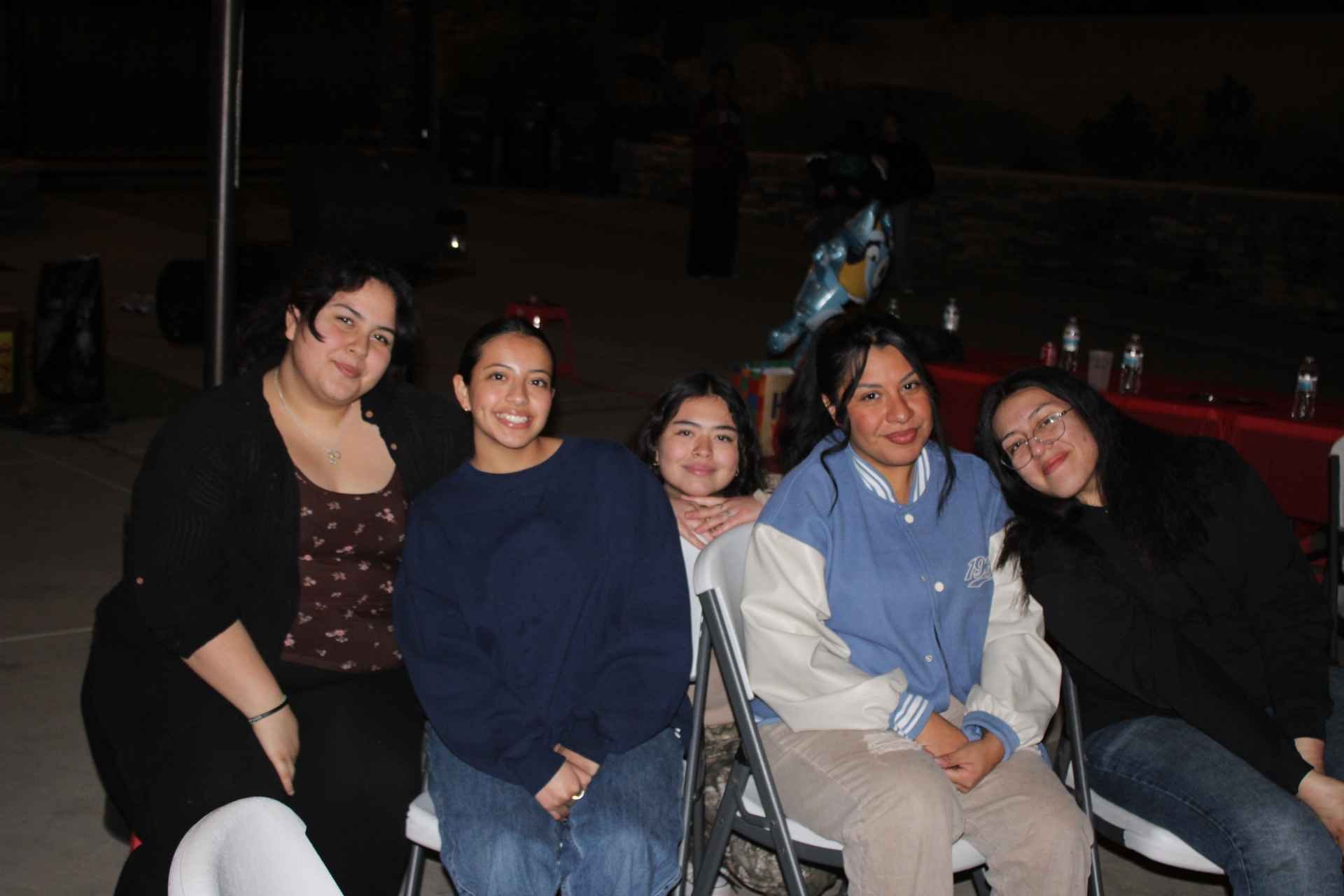 Group of five women sitting together at an outdoor event, smiling, with drinks and decorations in the background.