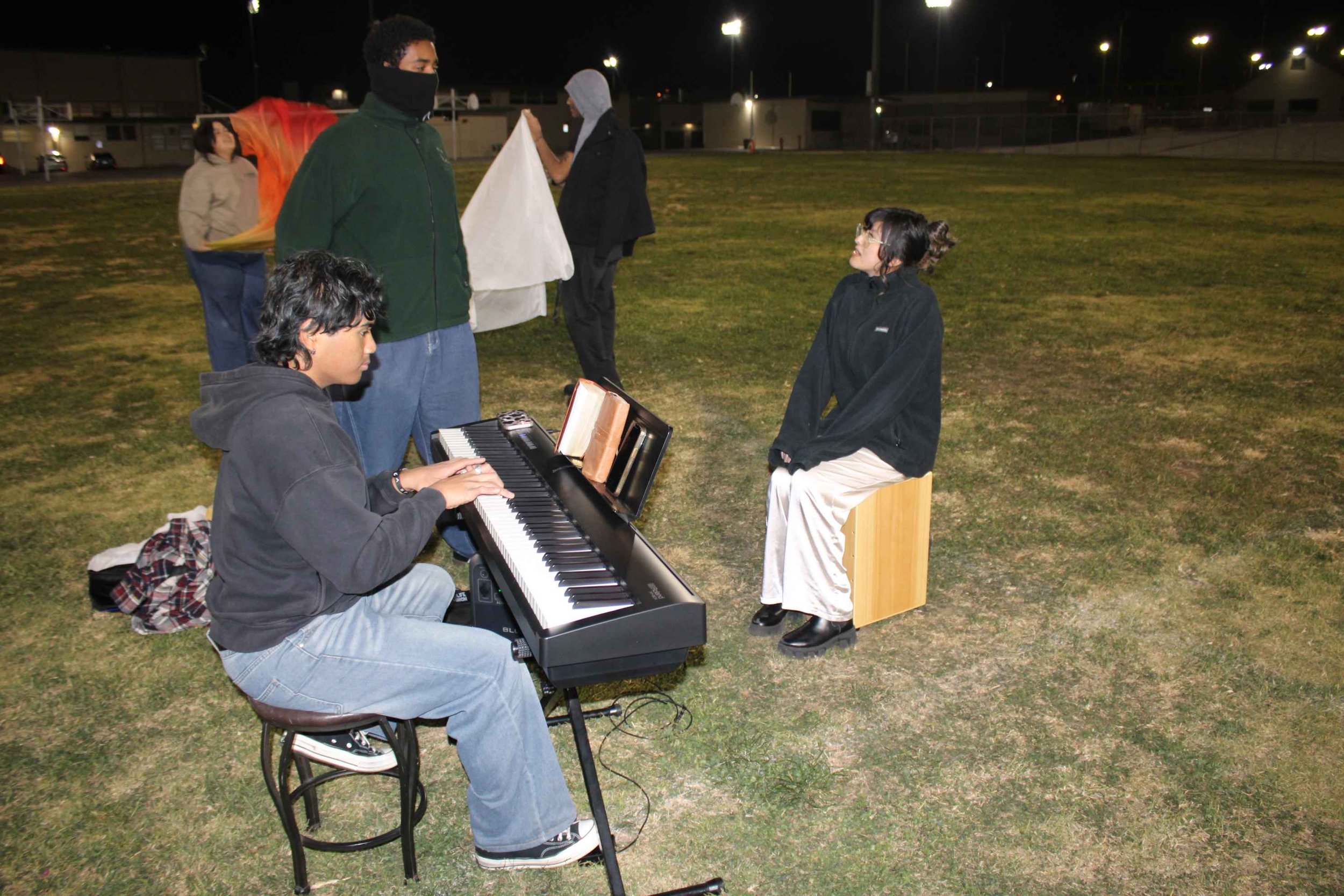 Group of people outdoors at night with a keyboard, some holding fabric, one person playing, another seated on a wooden box.