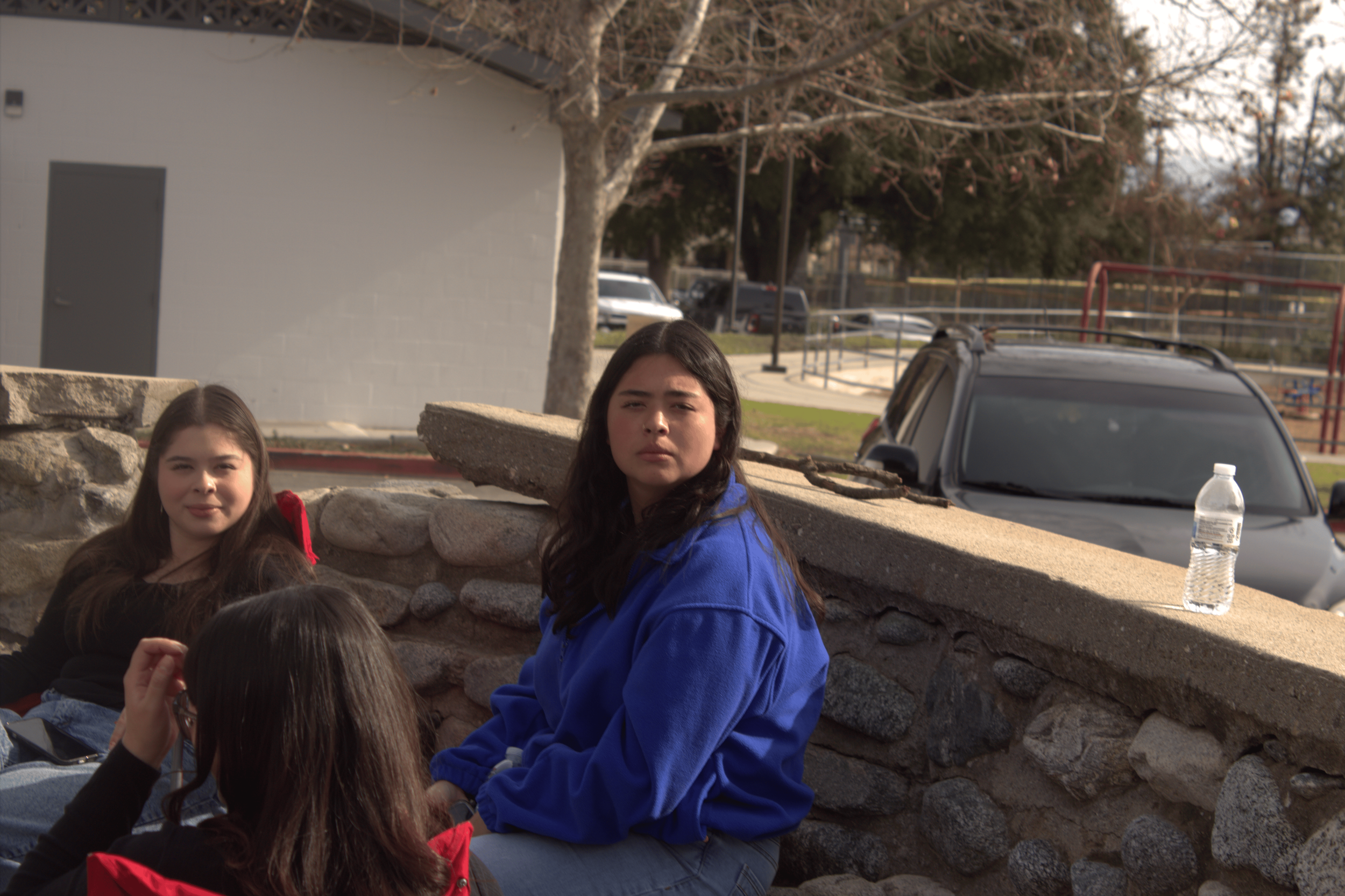 Three people sitting outdoors next to a stone wall, one wearing a blue jacket. A car and a plastic water bottle are visible in the background.