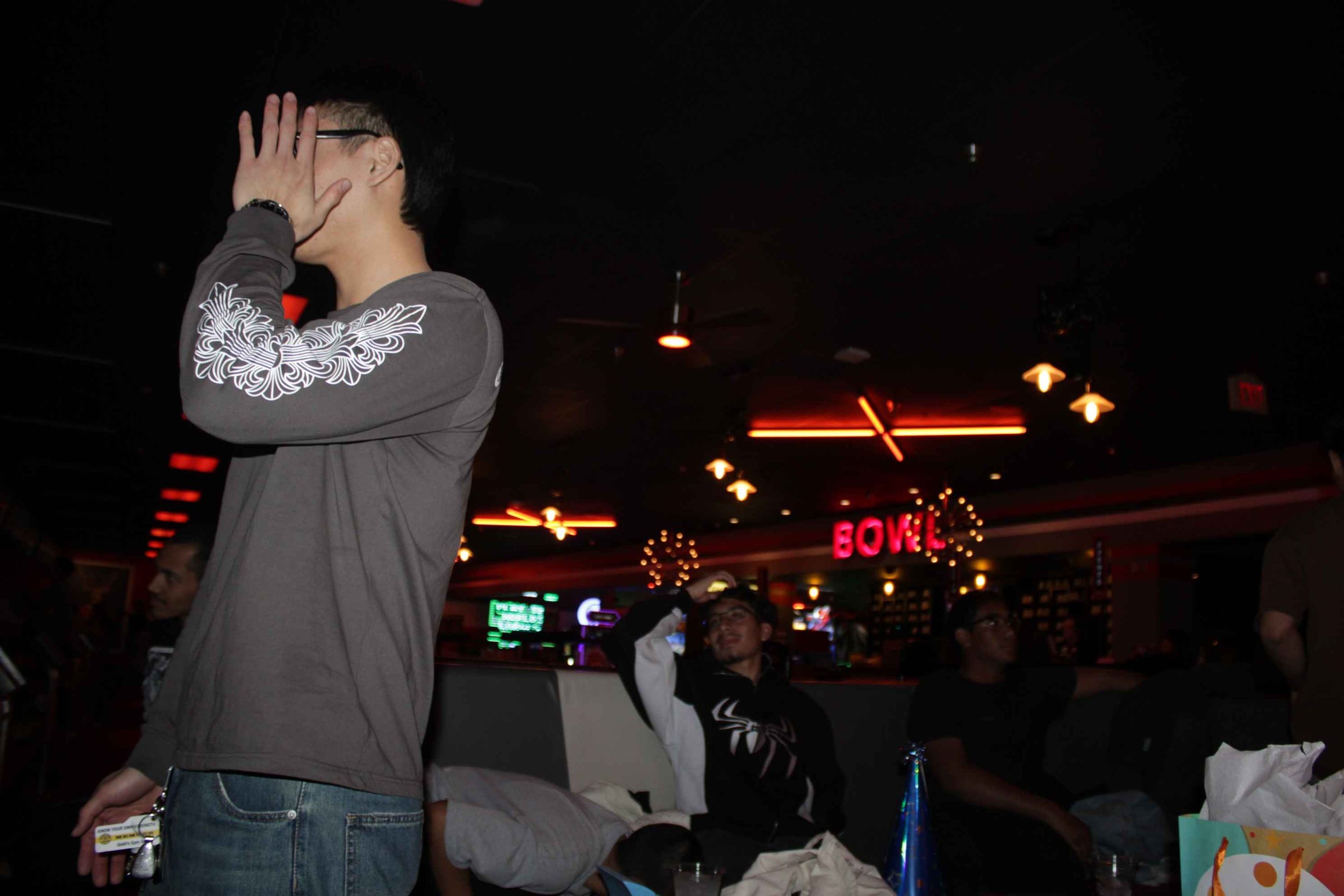 A person covering their face with one hand while standing in a dimly lit bowling alley with neon lights and several people sitting in the background.