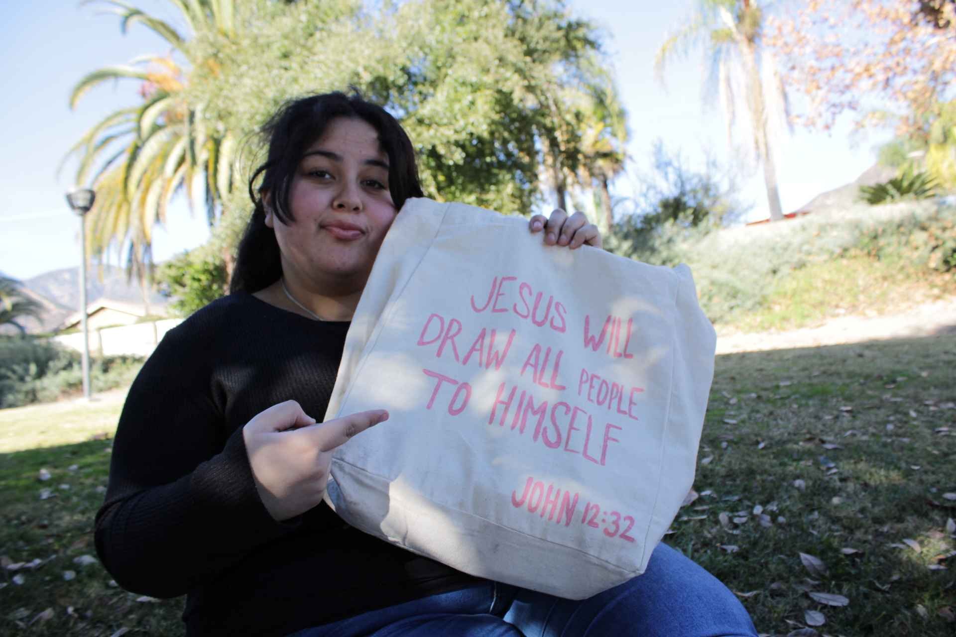 Person holding a tote bag with the text 'Jesus will draw all people to himself' and 'John 12:32' printed on it in a park setting.