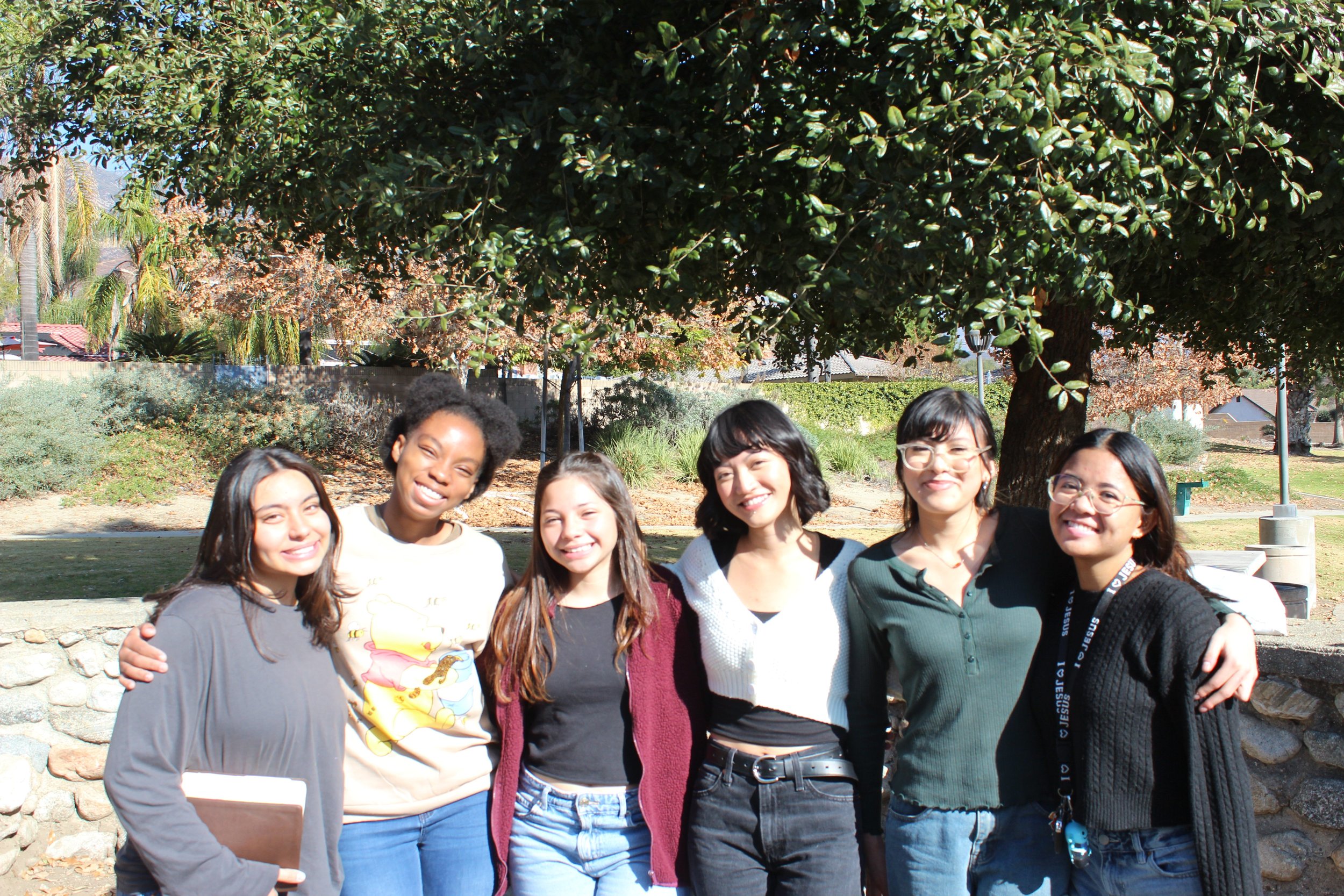Group of six young adults smiling and standing together in a park, with trees in the background. They are dressed casually, and one person holds a book.
