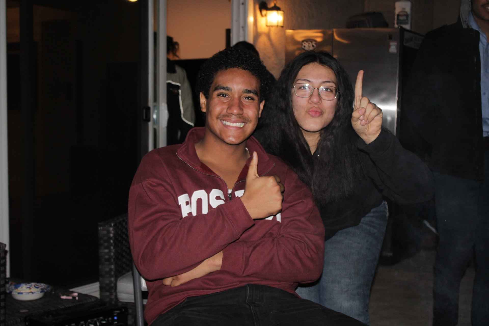 Two people posing indoors; one person is smiling and giving a thumbs-up while sitting, the other is making a funny face and pointing upward.