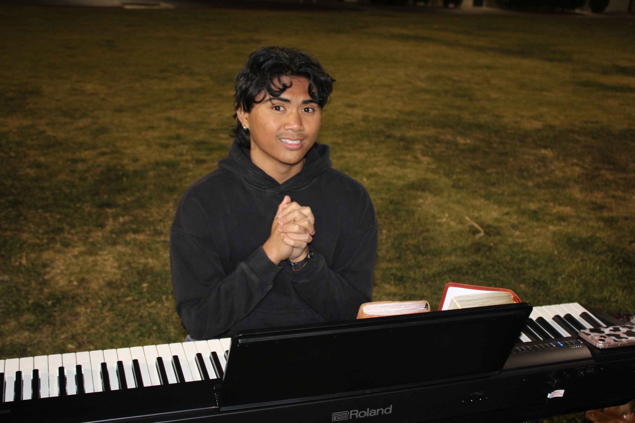 Person in black hoodie sitting at a Roland keyboard outdoors on grass.