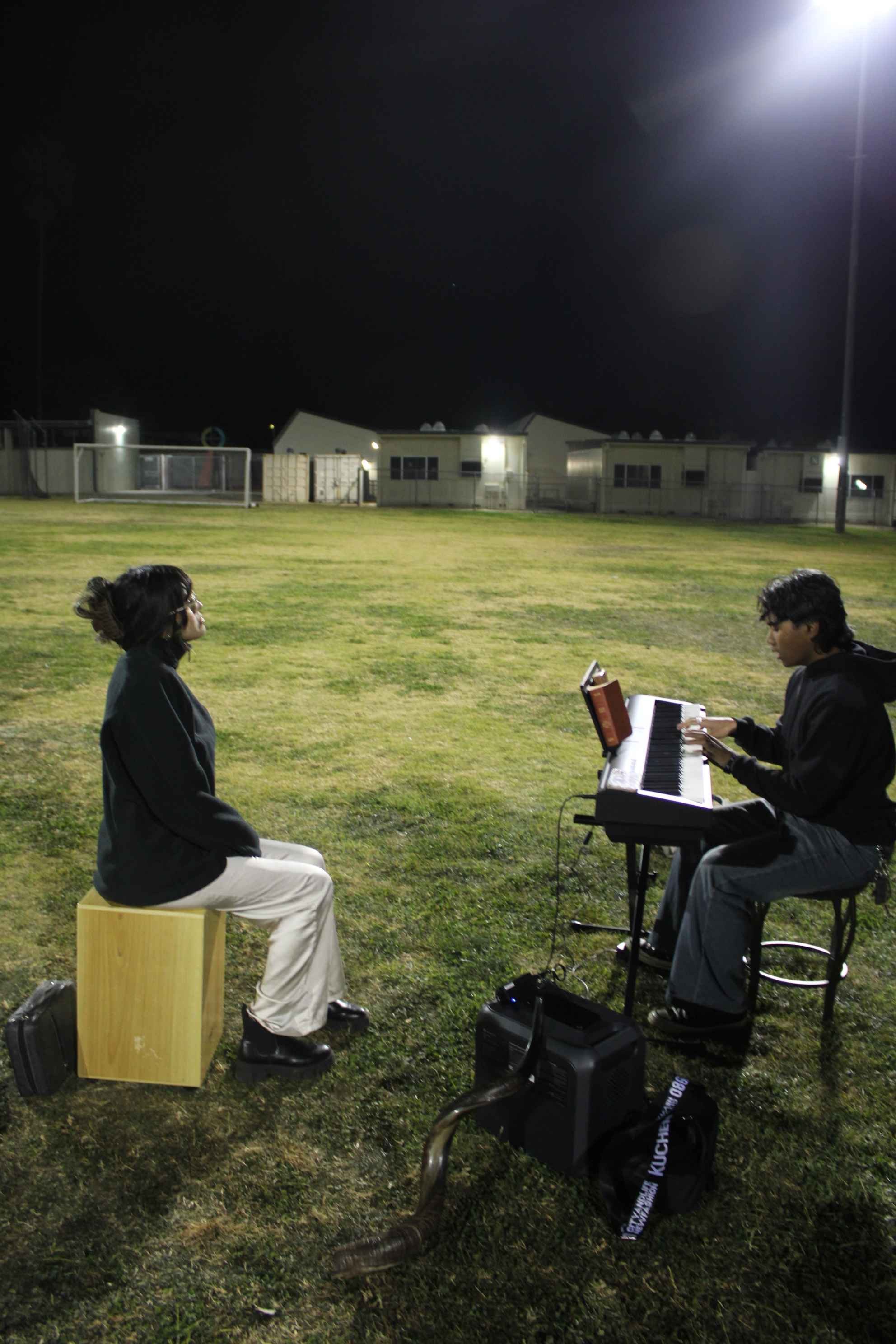 Two people outdoors at night, one playing a keyboard and the other sitting on a wooden box, with a portable speaker and cable nearby on grass.