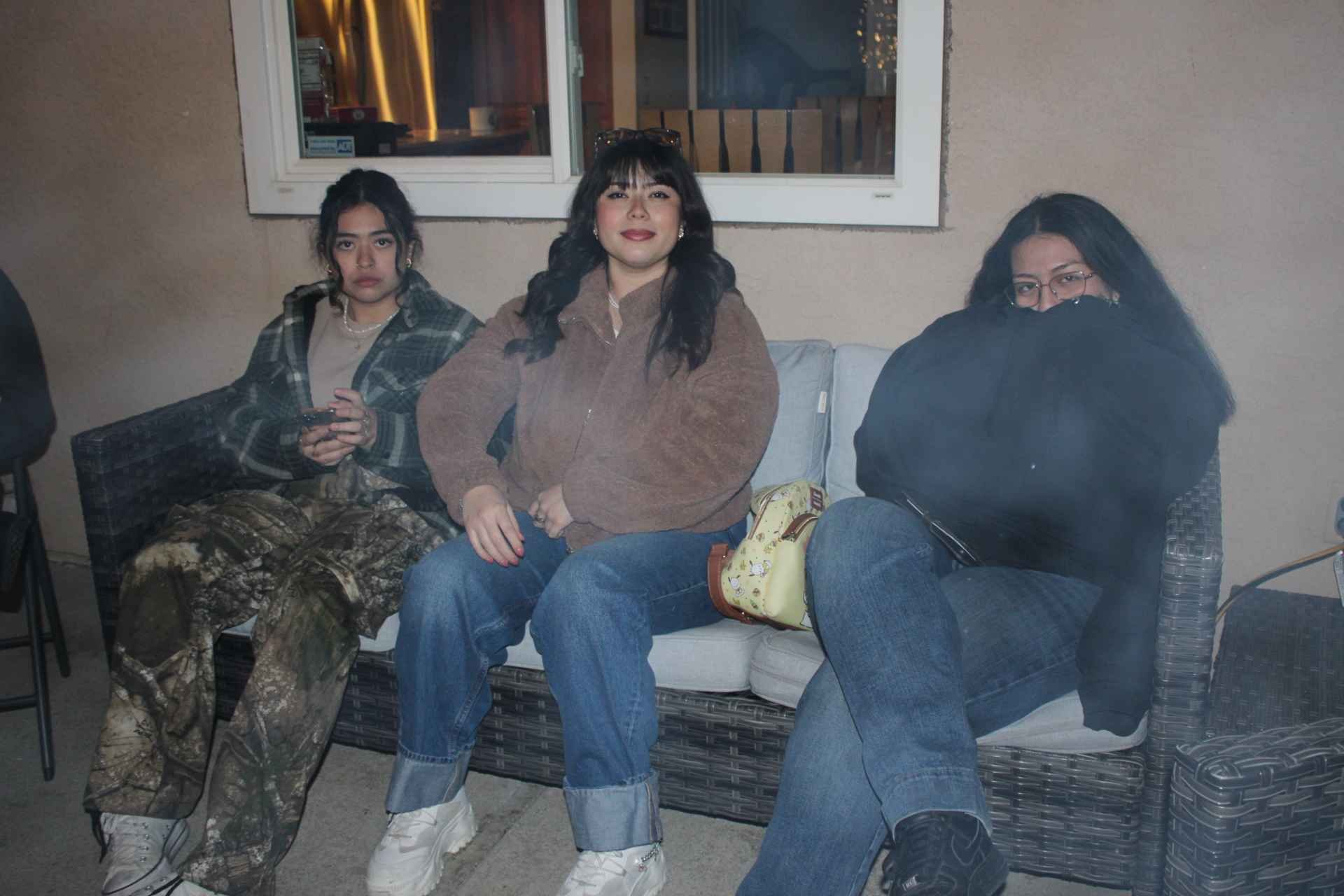 Three people sit on a wicker couch outdoors at night, wearing casual winter clothing.