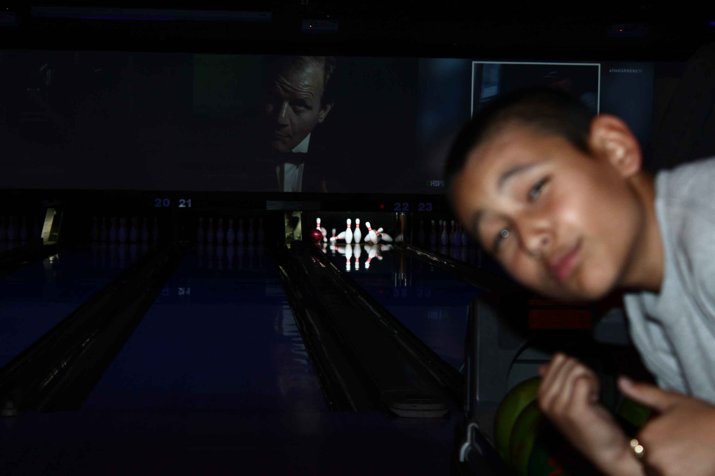 Boy posing in a bowling alley with lanes and pins visible; a large screen in the background shows a movie.