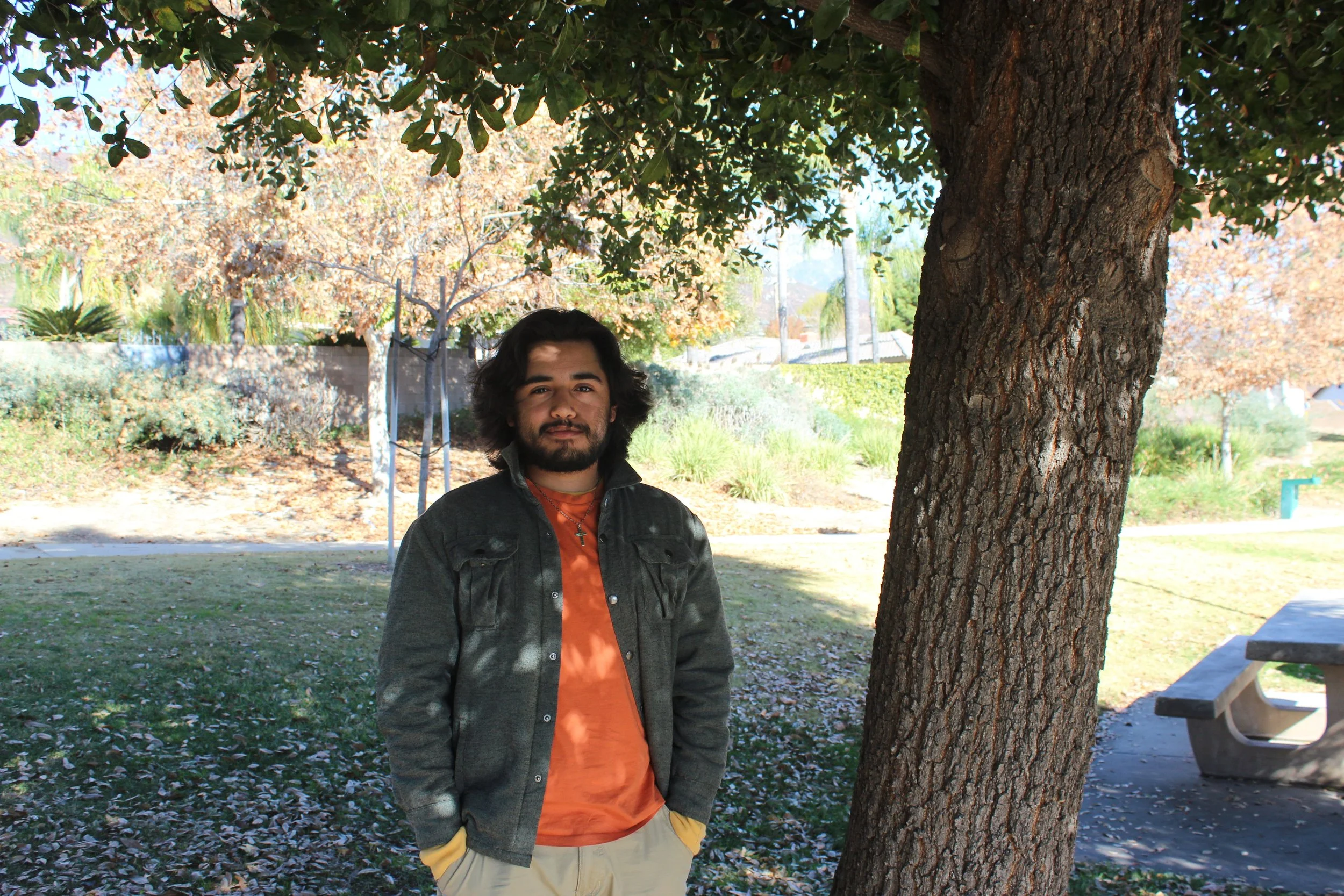 A person standing under a tree in a park setting, wearing an orange shirt and gray jacket, with hands in pockets. The background shows trees and a picnic table.