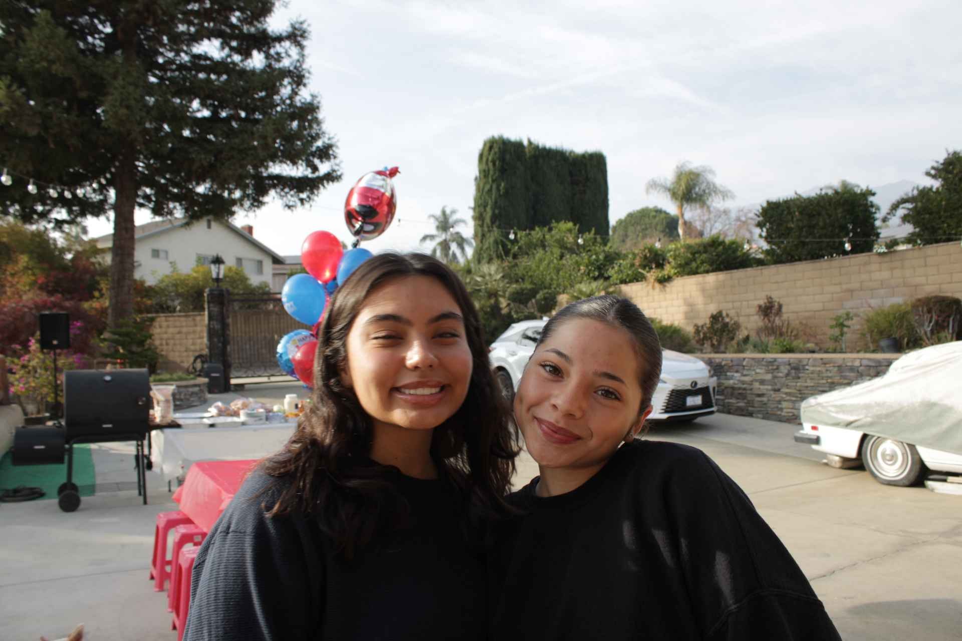 Two women smiling in a backyard with red and blue balloons and a white car in the background.