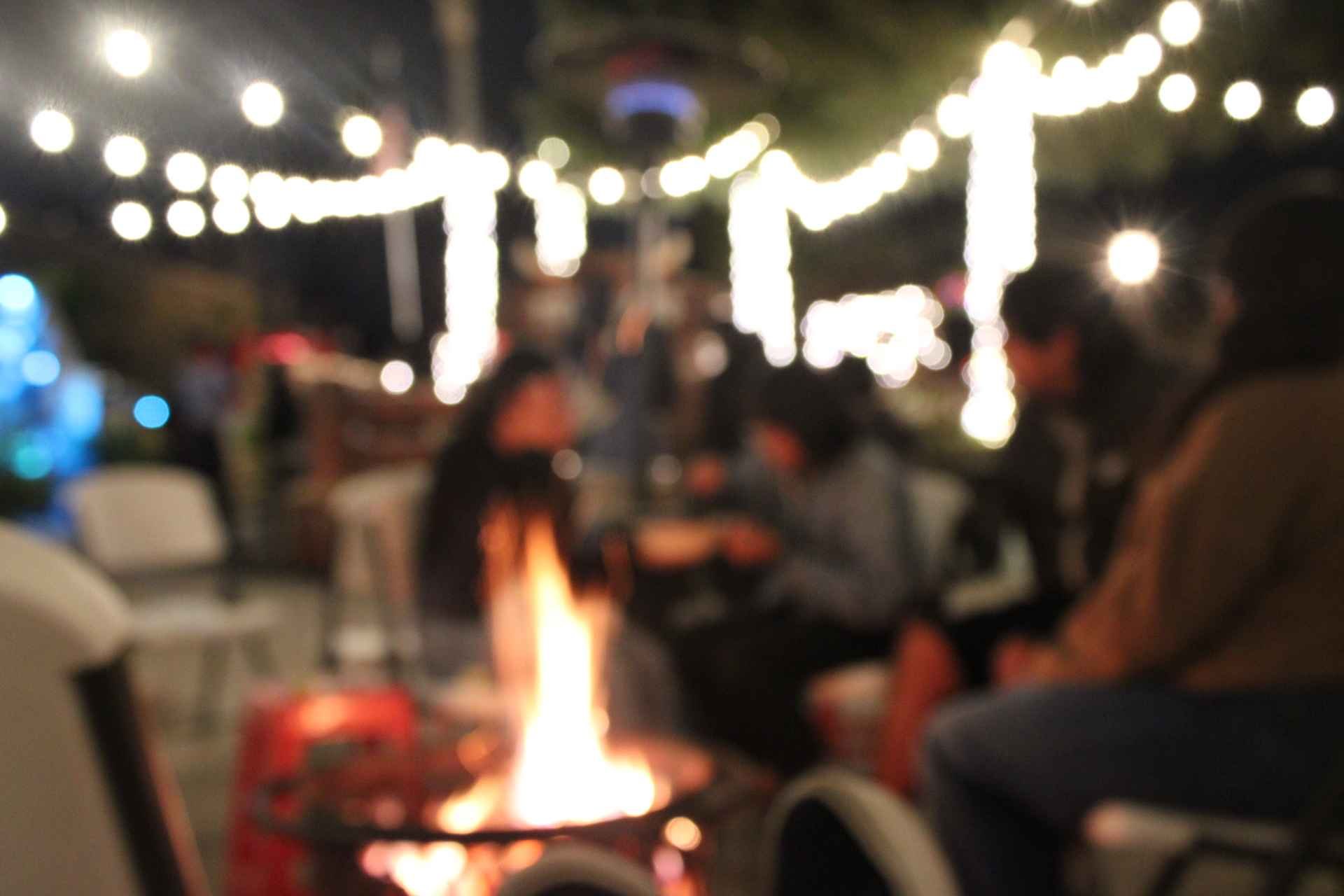 Blurry image of an outdoor gathering at night with string lights and people around a fire pit.