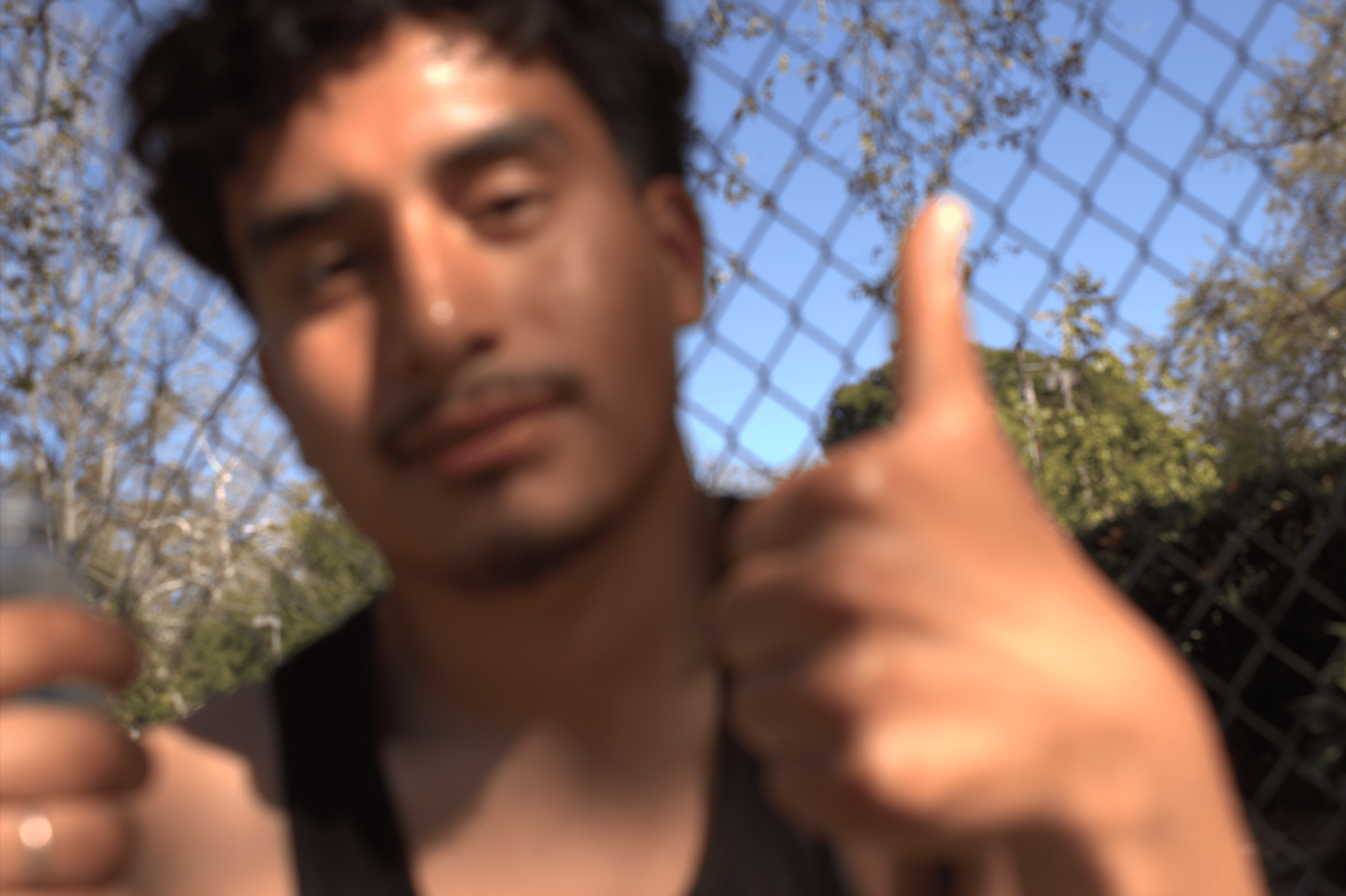 Blurred person giving a thumbs-up gesture in front of a chain-link fence with trees and blue sky in the background.
