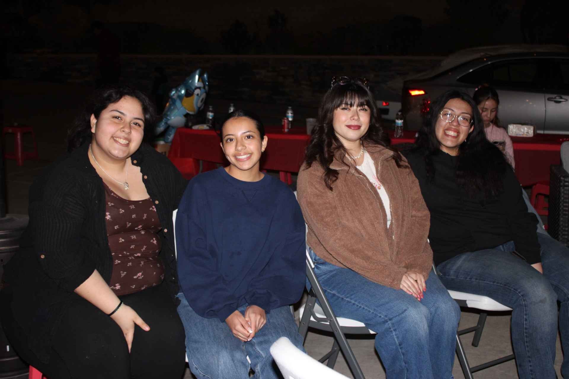 Four people sitting together in chairs, smiling at the camera, with a car and a decorated table in the background at night.