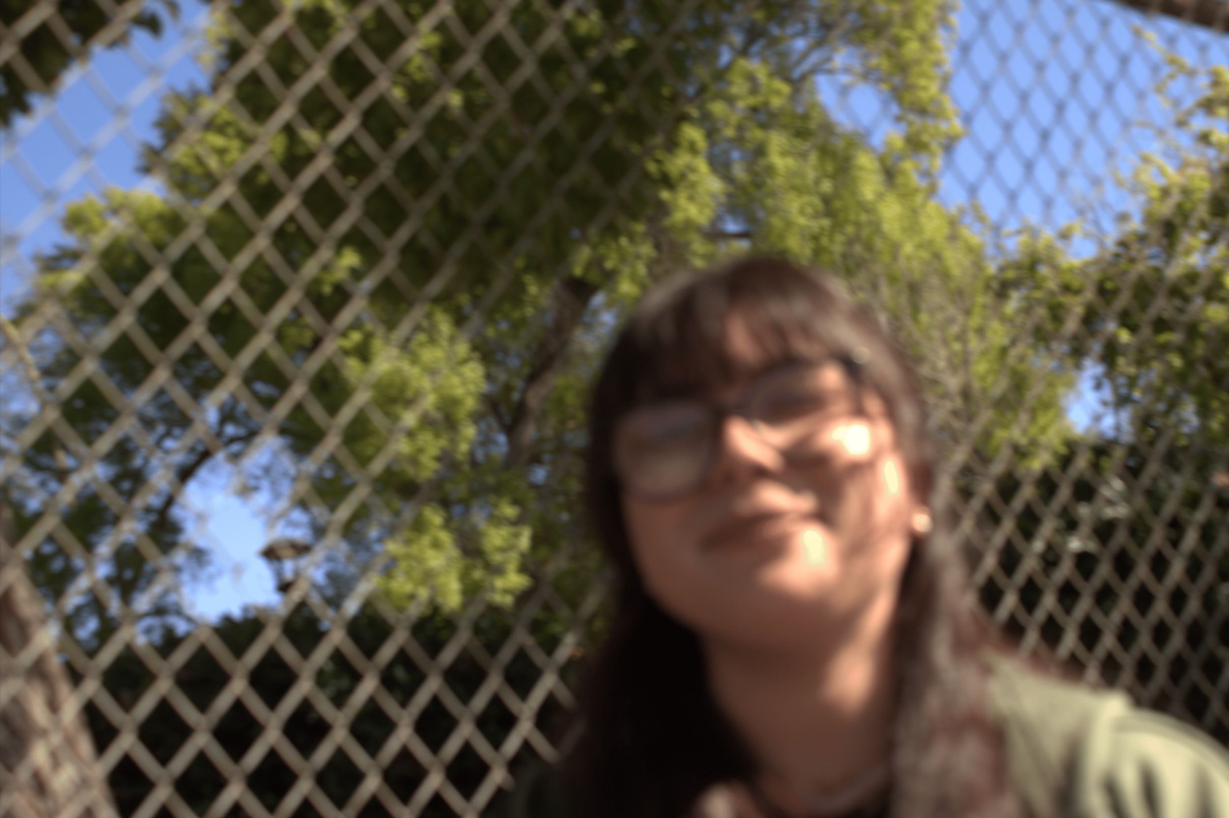 Blurry photo of a person in front of a chain-link fence with trees and blue sky in the background.