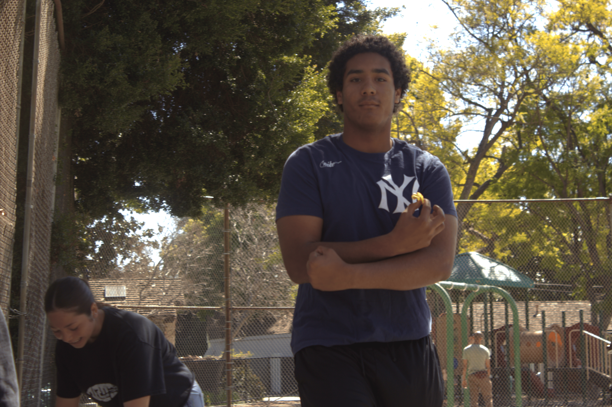 Person in a blue T-shirt standing outdoors by a playground, next to a woman bending down, with trees and a fence in the background.