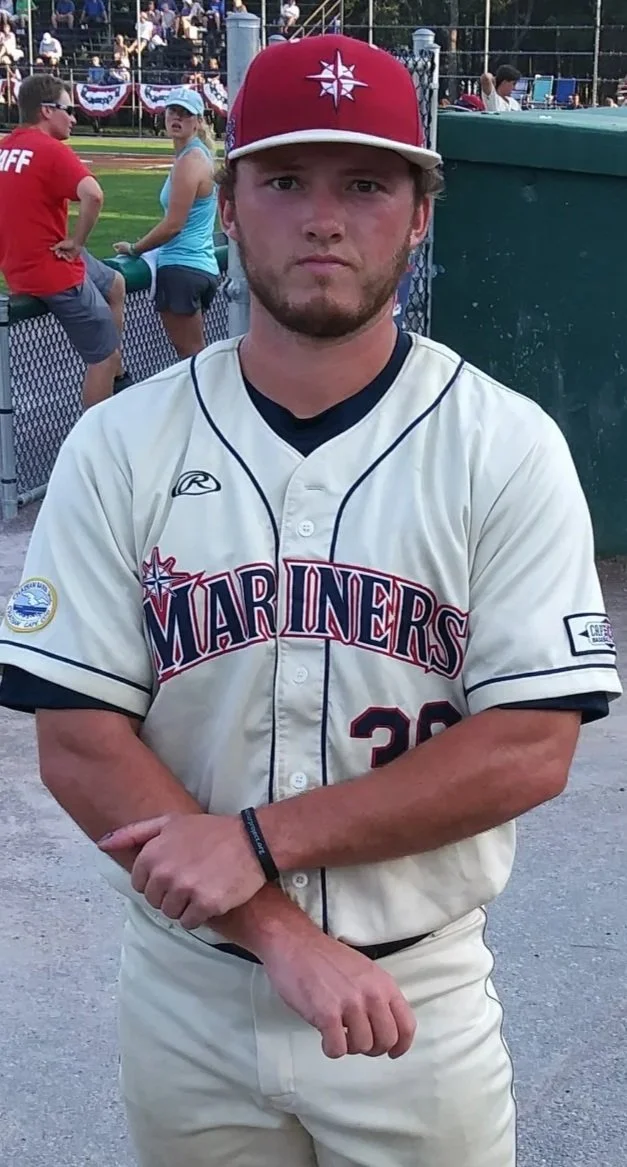 A young man with light skin and brown hair, wearing a Mariners baseball jersey and a red cap, standing with his arms crossed at a baseball game.