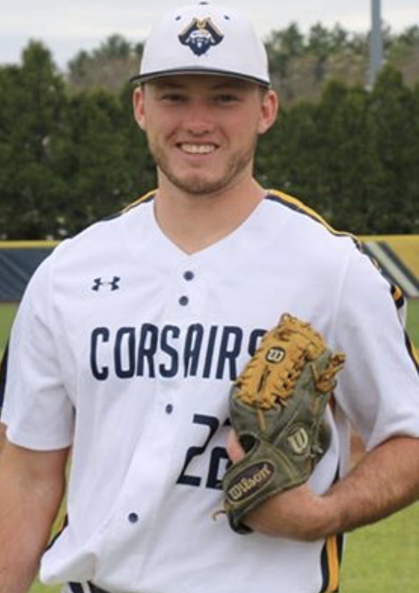 A young man wearing a baseball uniform and cap with a beard, smiling, standing on a baseball field holding a baseball glove.