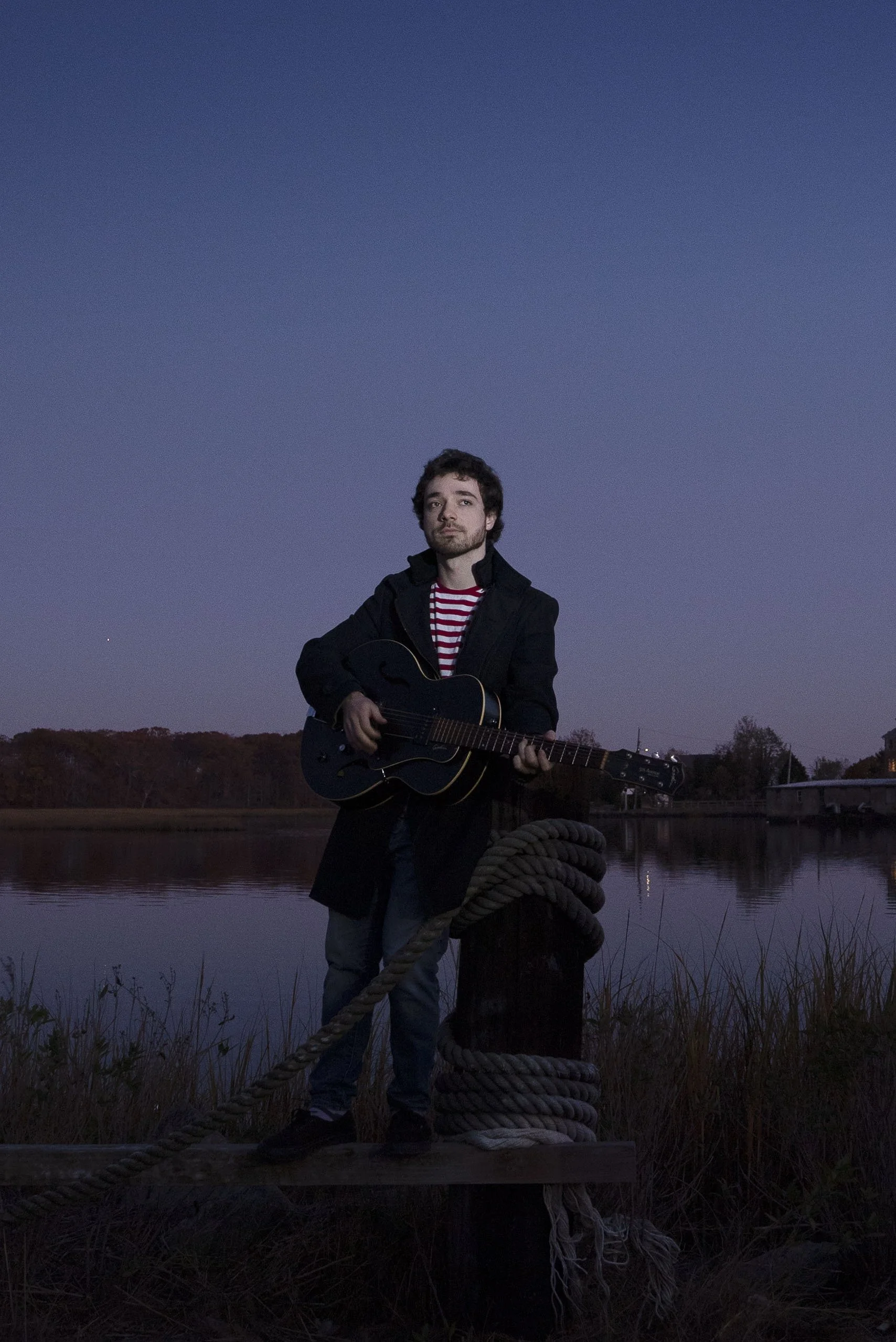 A man standing on a dock by a body of water at dusk, holding a guitar. He is wearing a dark coat and a red and white striped shirt, with trees and a bridge visible in the background.