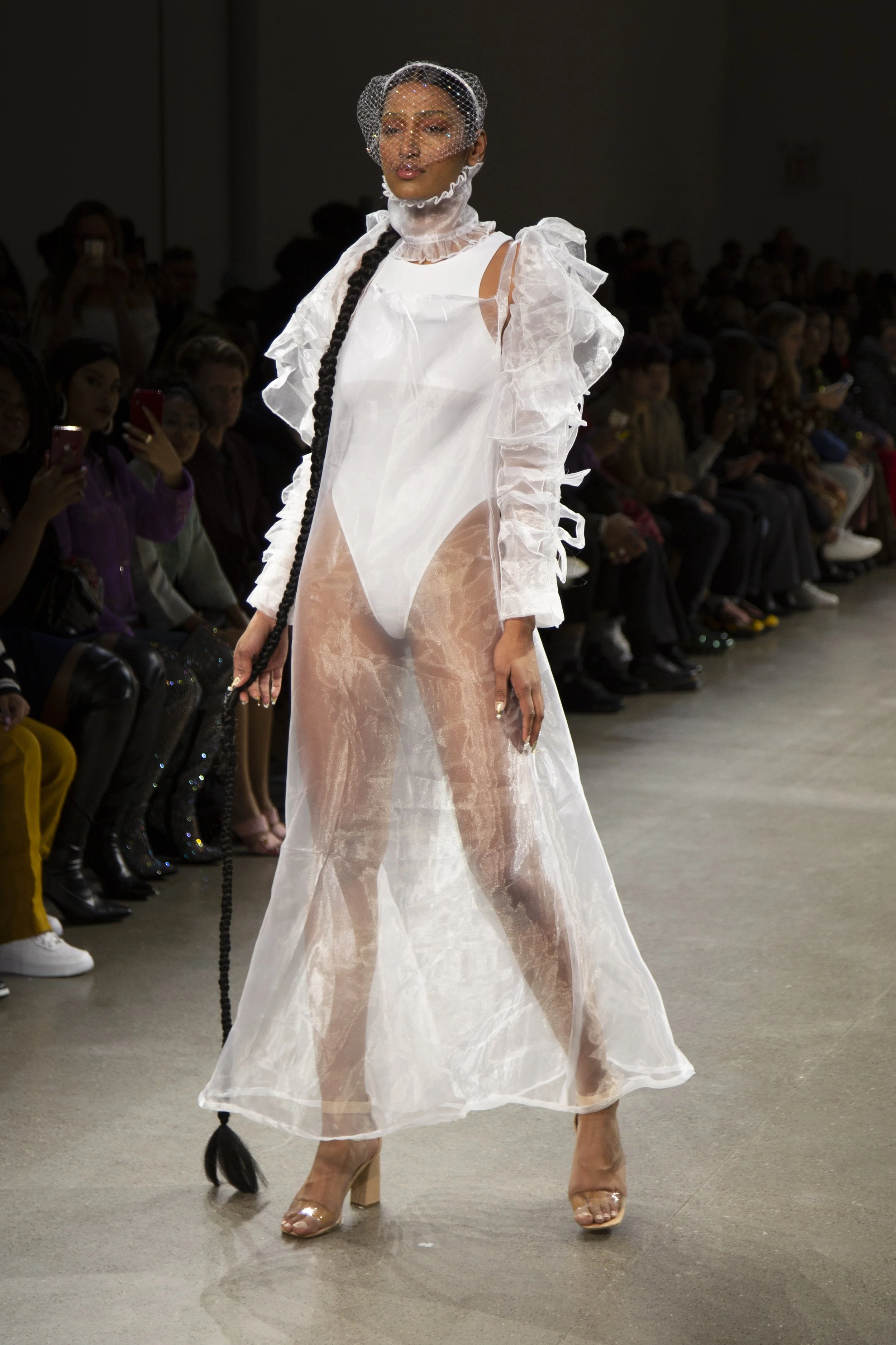 A model walking on a runway wearing a sheer, ruffled white dress with gloves, open-toe heels, and a birdcage veil, with an audience watching in the background.