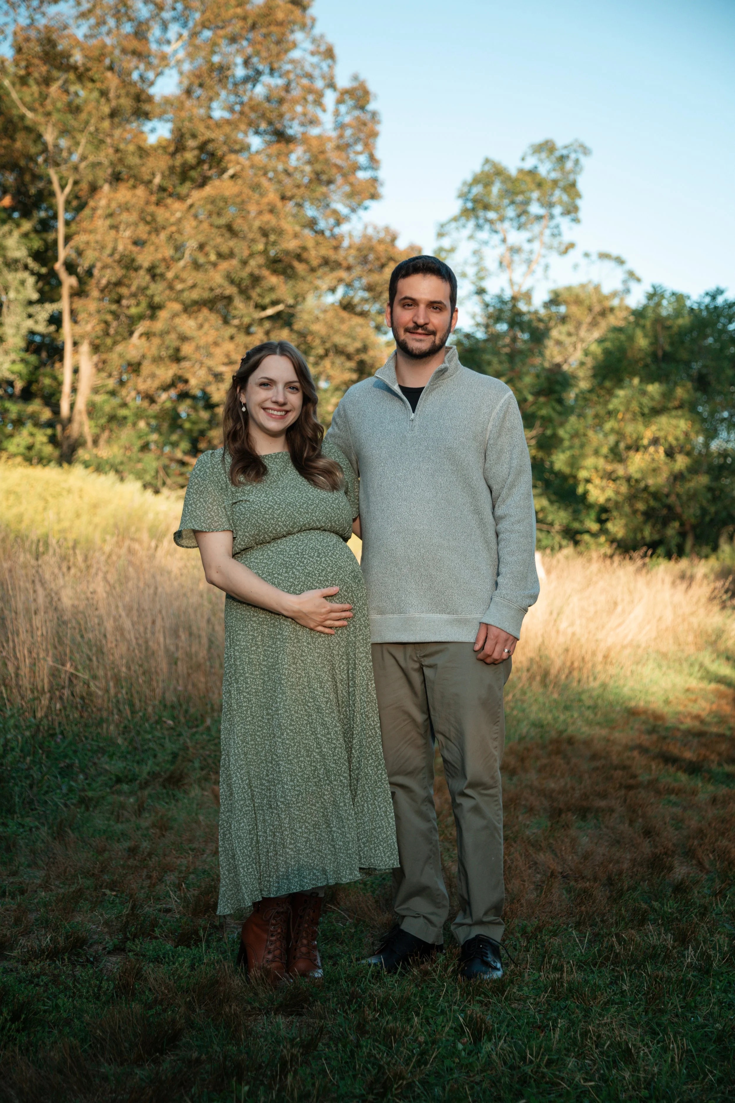 A pregnant woman in a green dress and brown boots stands next to a man in a gray sweater and khaki pants, both smiling outdoors with trees and grass background.