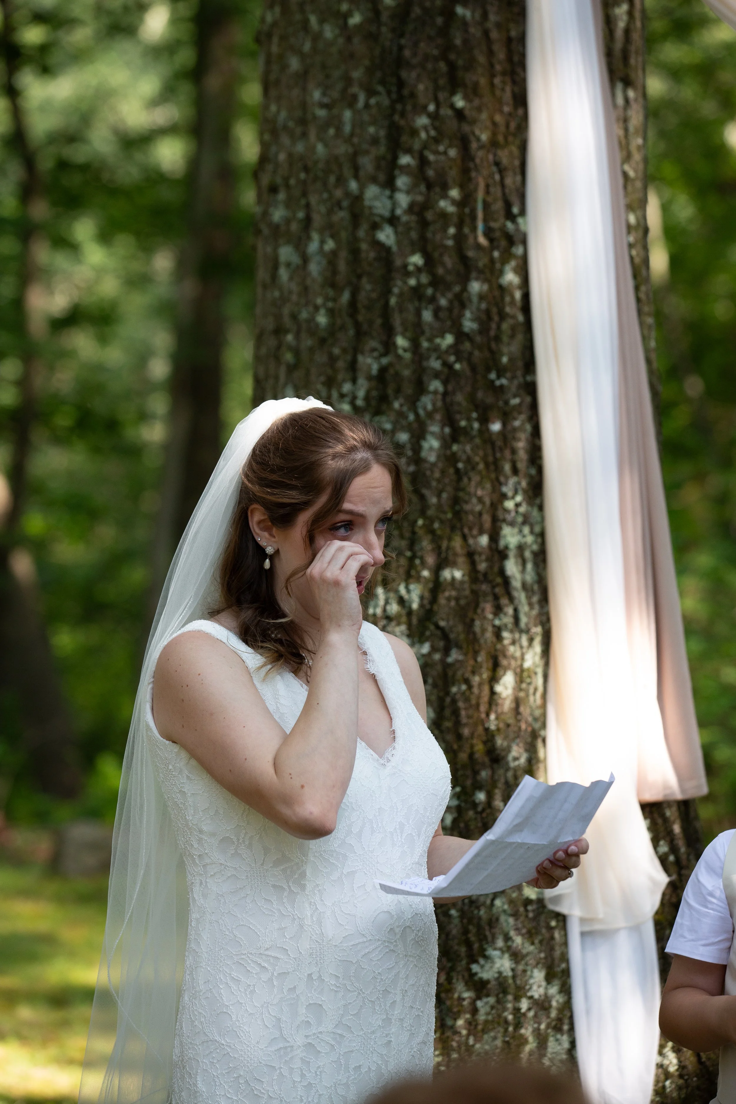A woman in a white wedding dress and veil is crying while holding a piece of paper during an outdoor wedding ceremony in a forest.