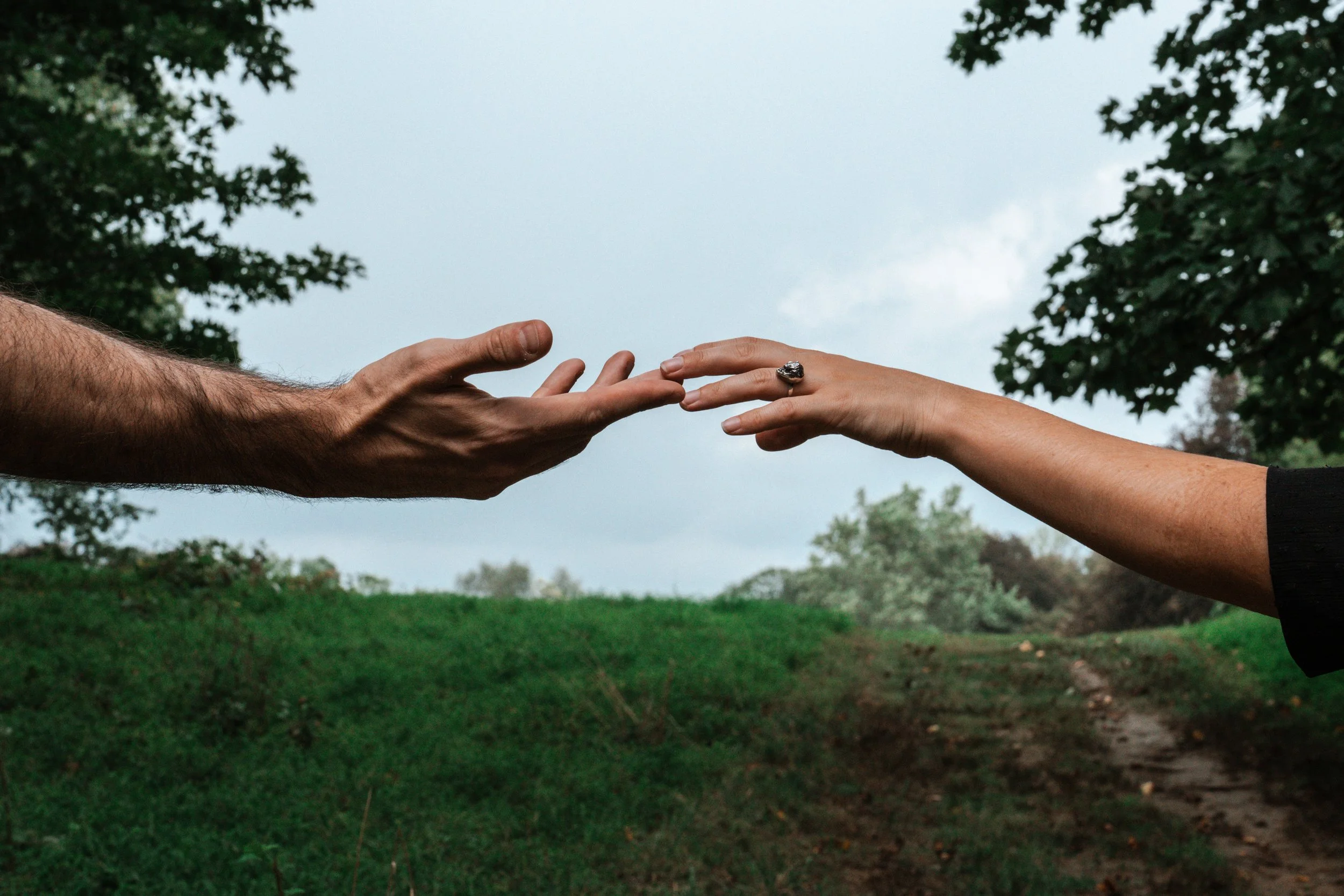 Two hands reaching towards each other outdoors with a cloudy sky and green trees in the background.