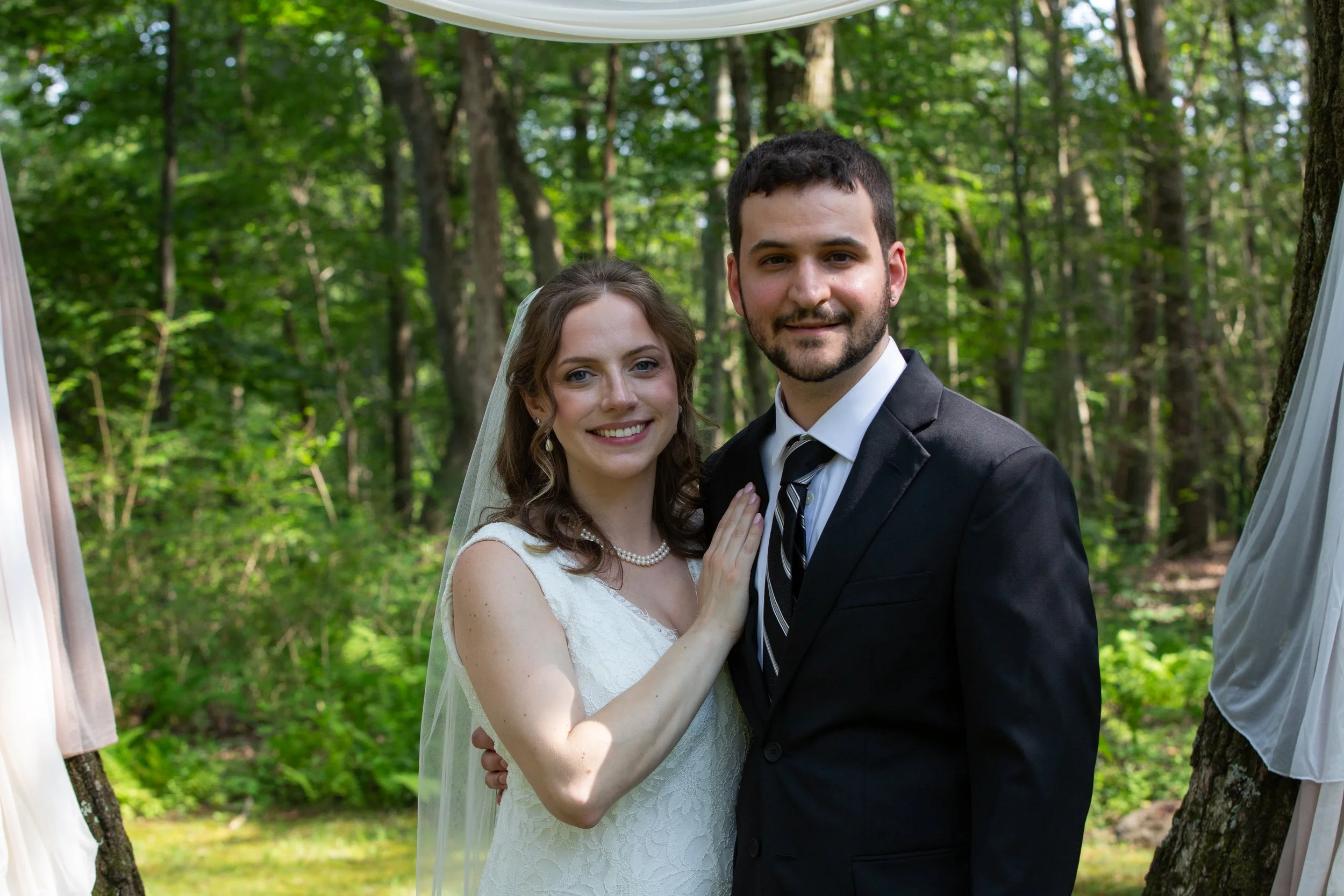 A bride and groom in wedding attire outdoors in a wooded area, smiling at the camera.