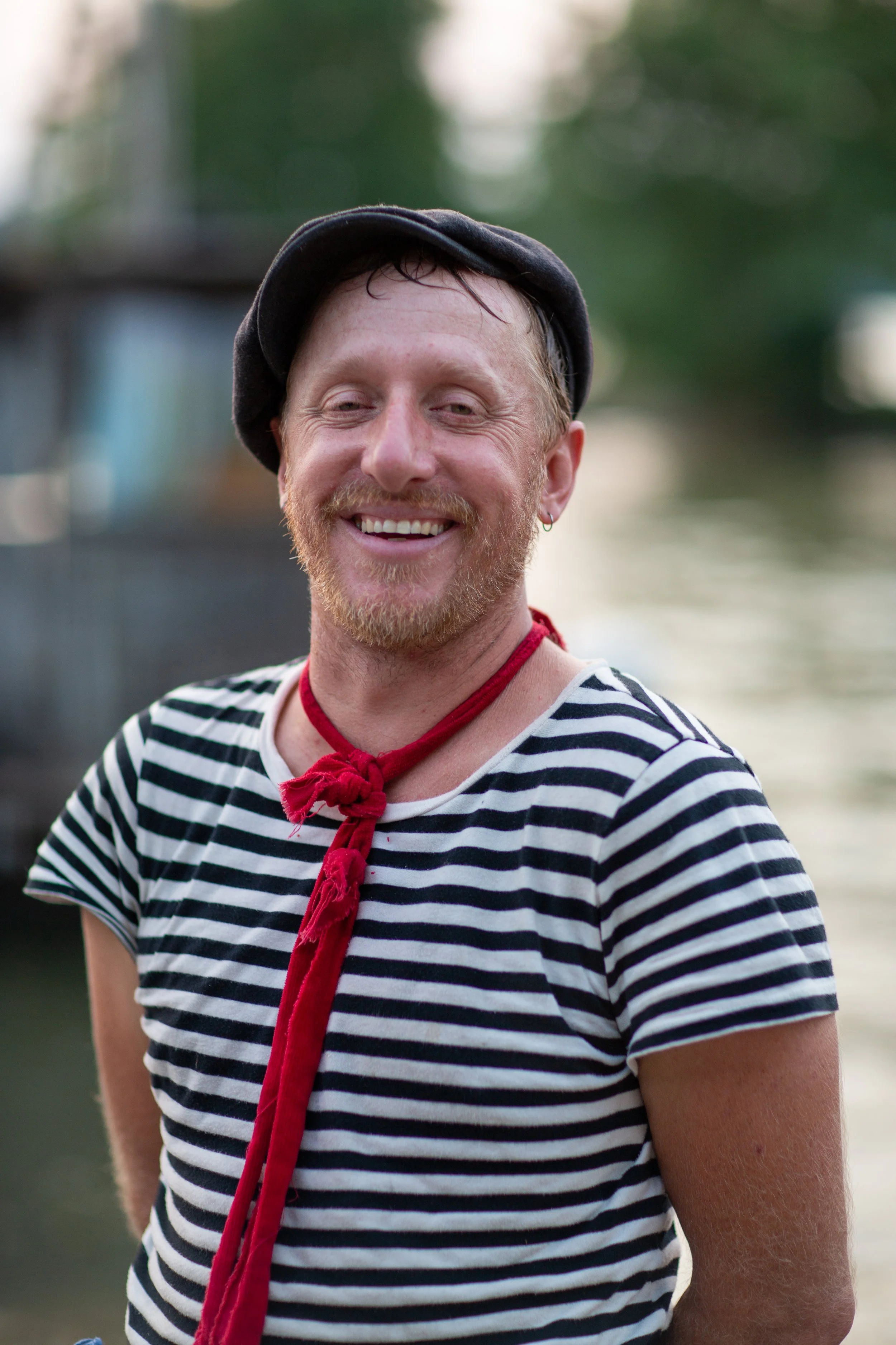 Smiling man with a red beard and mustache wearing a striped shirt, black beret, and red neck scarf, standing outdoors near water.