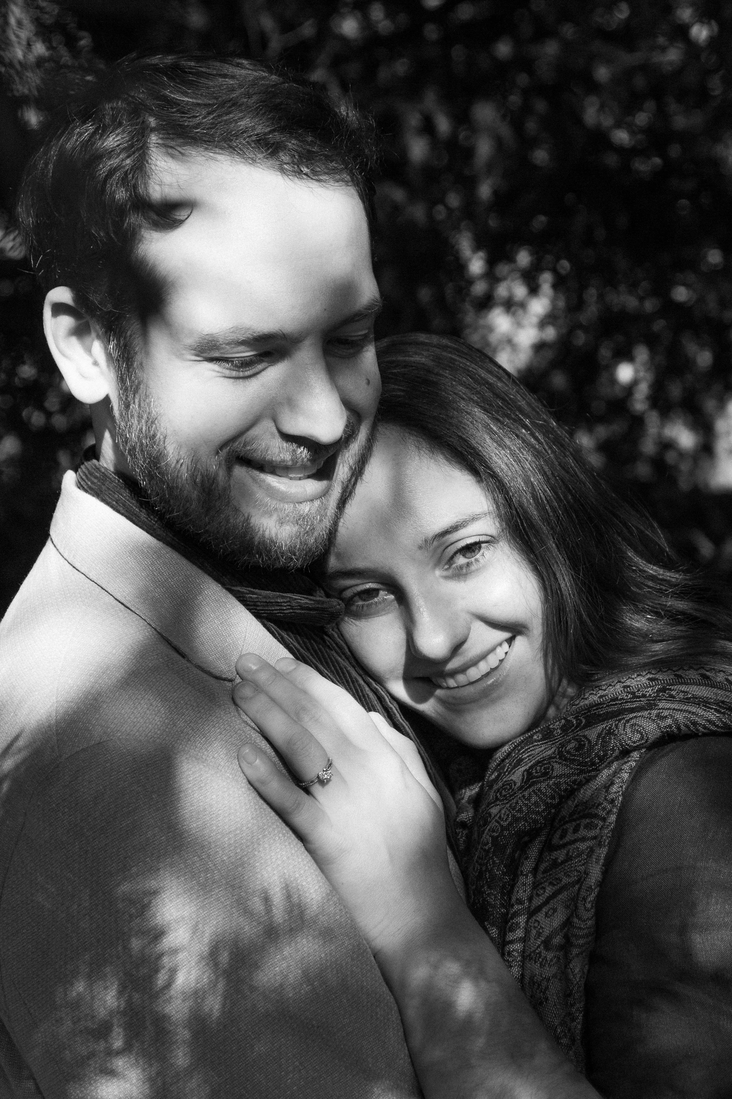 A black and white photo of a happy couple embracing tightly outdoors, with the woman smiling and showing an engagement ring on her finger.