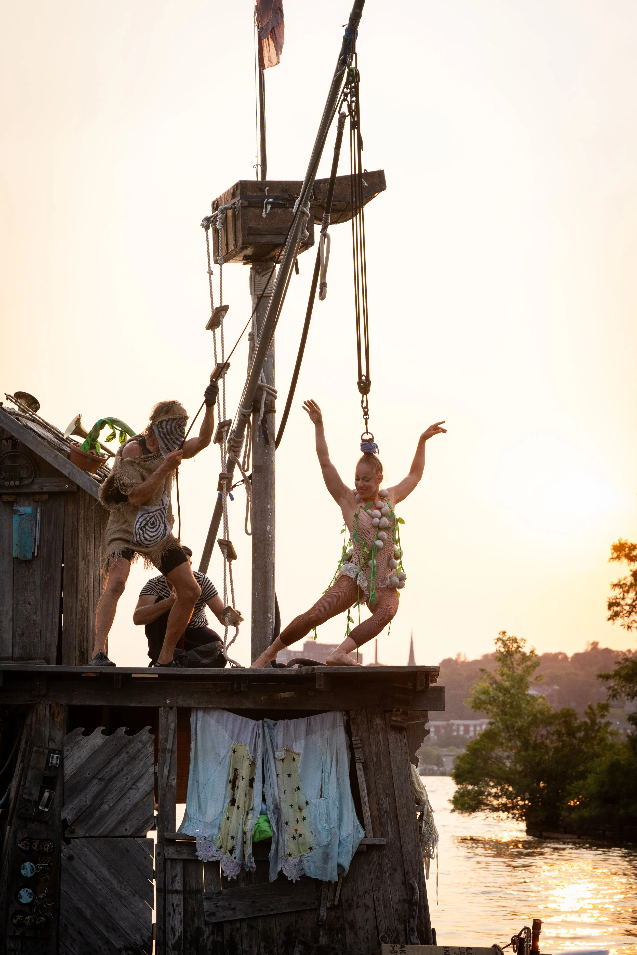 A performer in an elaborate costume with floral and pom-pom decorations is on a swing, performing a pose on a wooden platform at sunset, with crew members assisting.