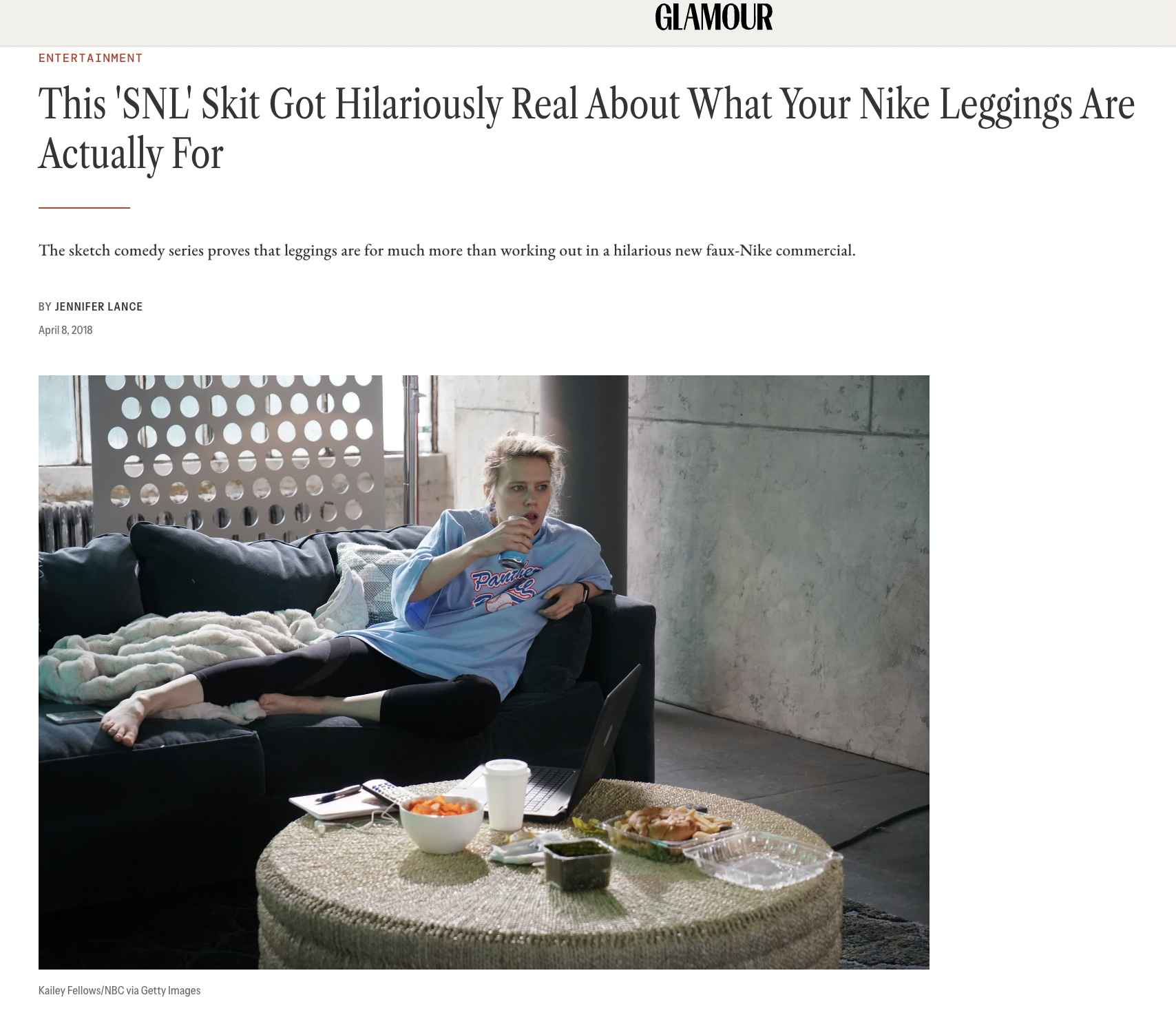 A woman sitting on a couch in a casual living room, eating food from a container with a laptop and additional food containers on a round table in front of her.