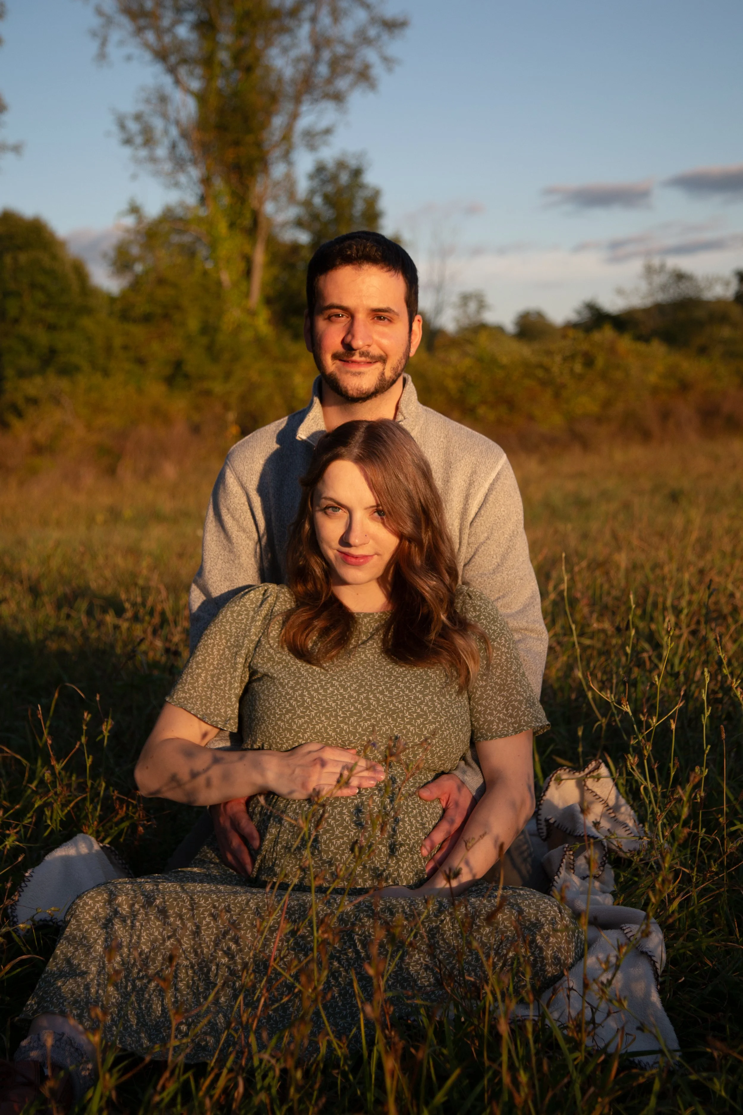 A pregnant woman and a man sitting outdoors in a field during sunset, the woman sitting on the ground with the man kneeling behind her, both smiling at the camera.
