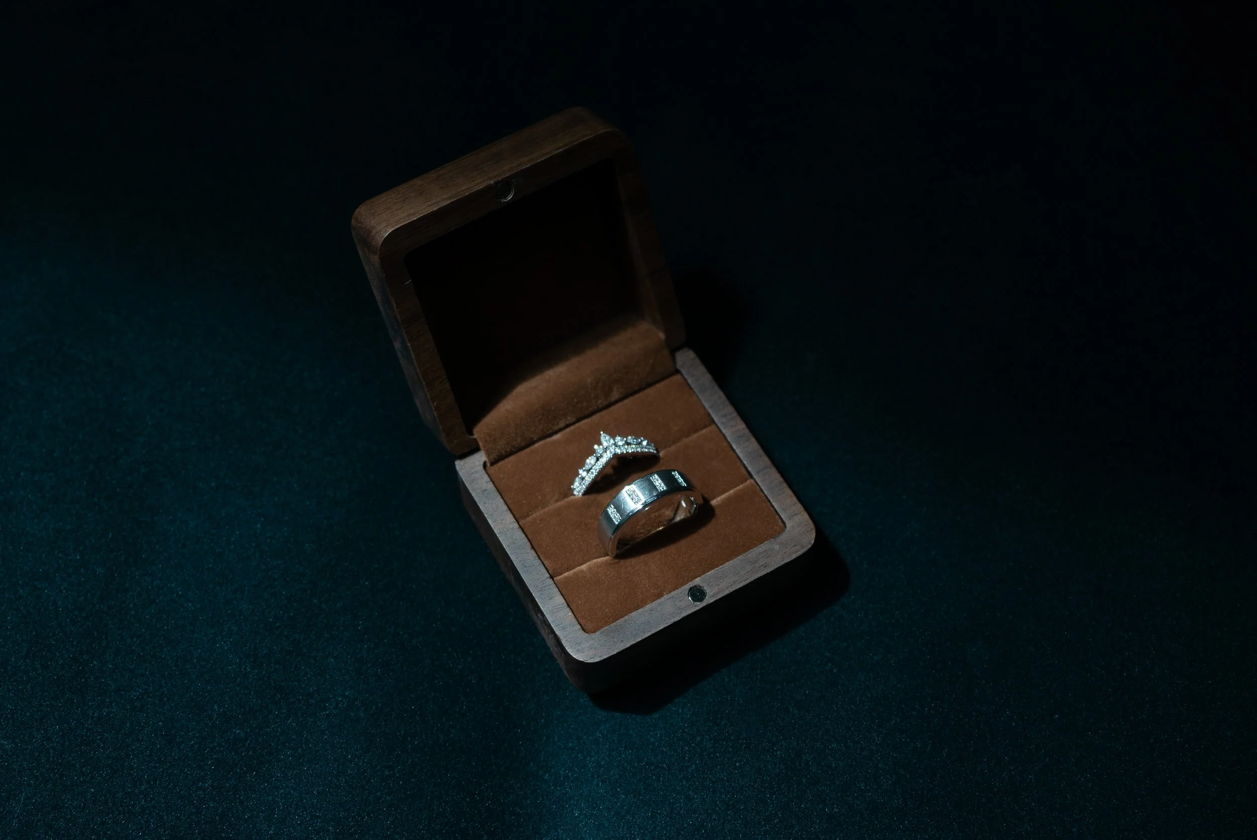 A wooden jewelry box open to reveal two silver rings, one with diamonds, on a dark surface.
