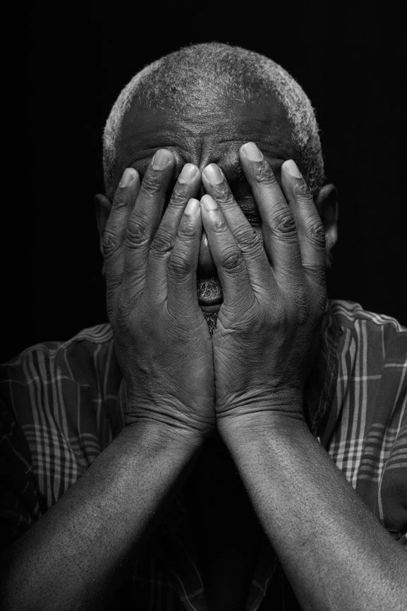 A black and white portrait of an elderly man with short hair covering his face with both hands, showing wrinkles and signs of age.