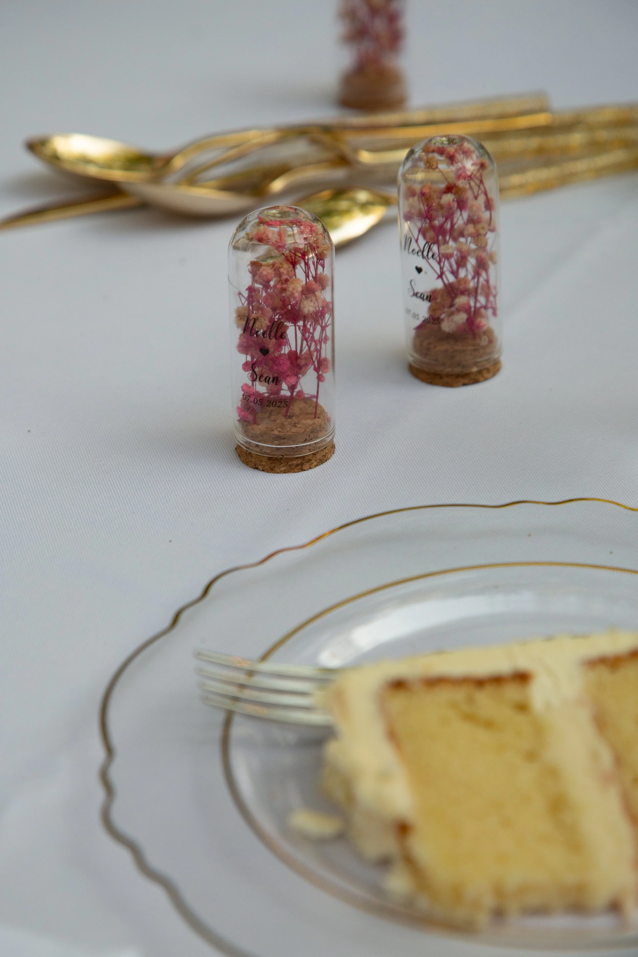 Two small glass containers with cork bases holding pink and white dried flowers, decorated with handwritten text and a date, placed on a dining table near a slice of cake and gold-colored cutlery.