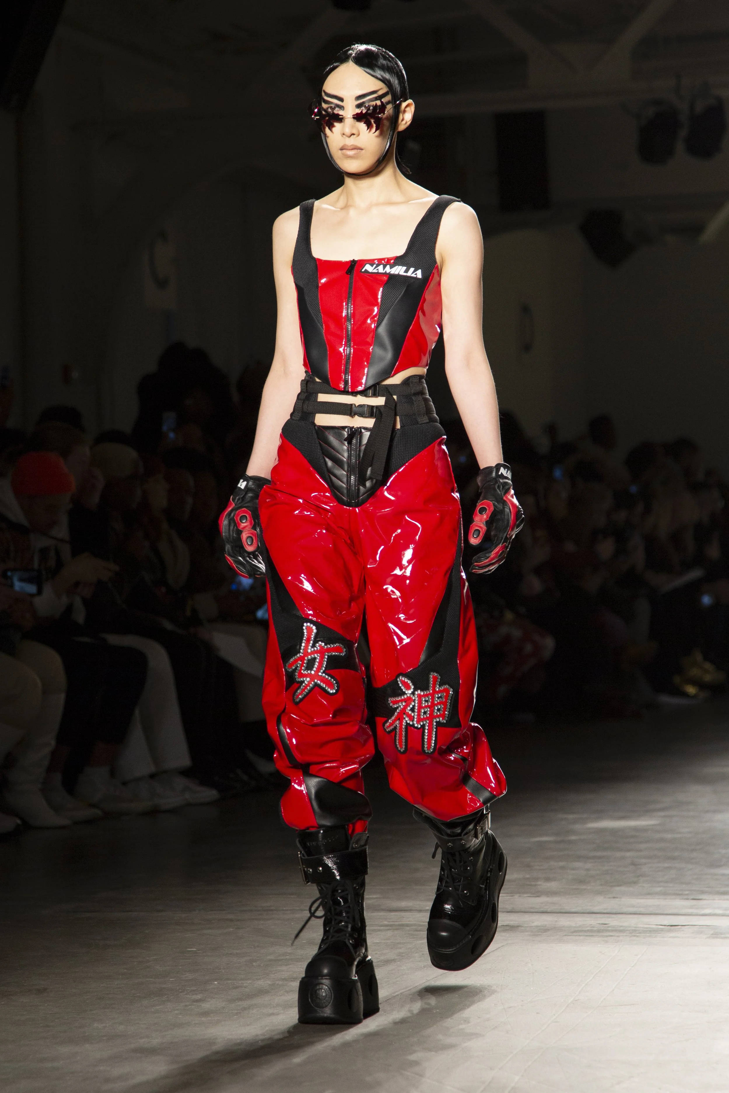 A fashion model walks down the runway in a red and black futuristic outfit with Japanese characters on the pants, wearing high-heeled boots and unique glasses, during a fashion show.