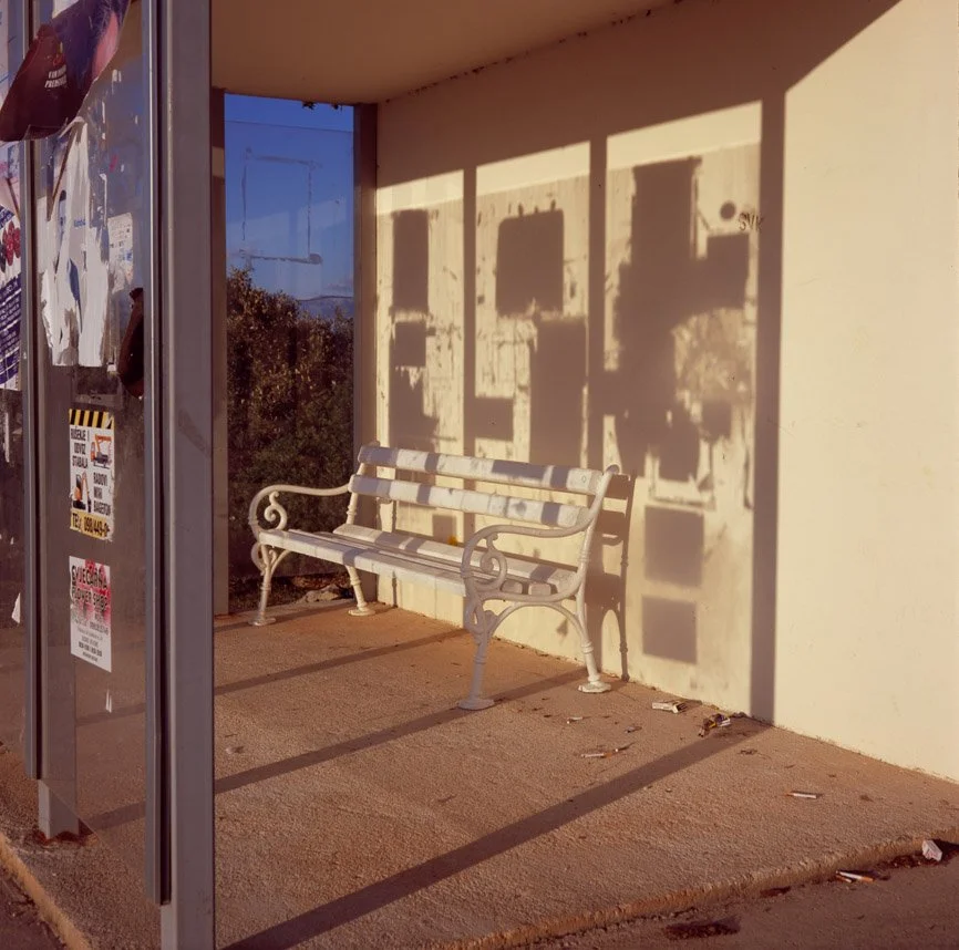 Empty outdoor bus stop shelter with a bench, cast shadows on the wall, and a nearby bulletin board with stickers and notices.