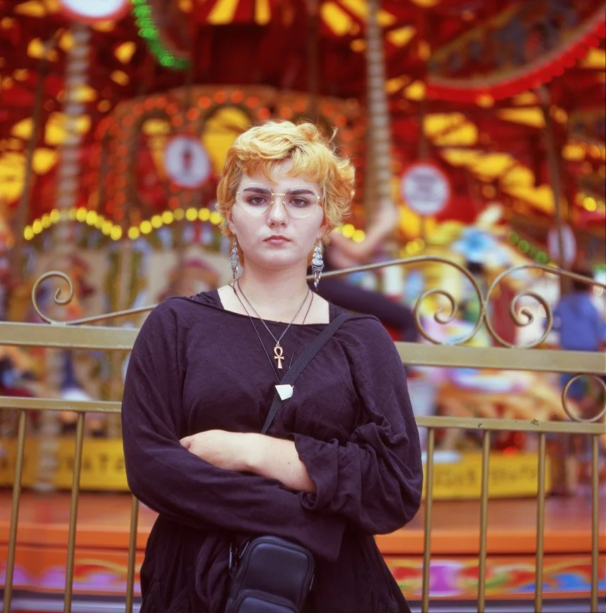 A woman with short blonde hair and glasses standing in front of a colorful carousel at an amusement park, wearing a black top and jewelry.